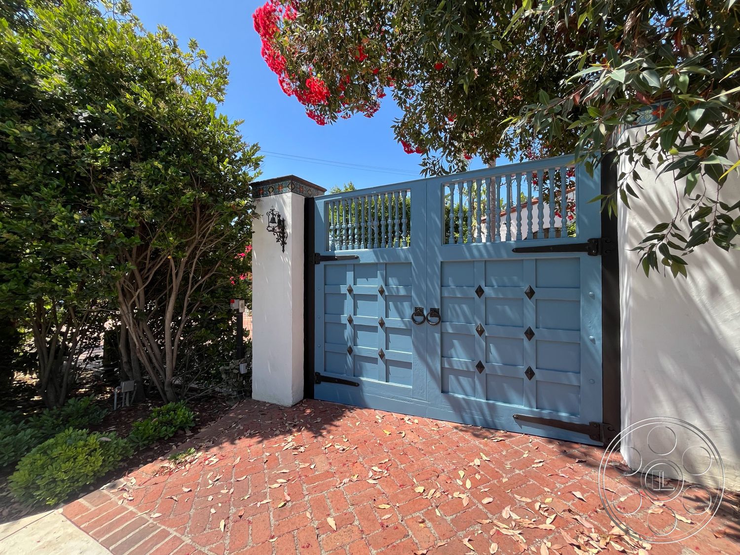 Medit 166 - exterior, brick driveway, dappled sunlight, white stucco wall, lush green landscaping, light blue wooden gate, ornate black lantern lighting, textured foliage accents, decorative vertical slats on gate, diamond-shaped hardware details, boundary garden setting, leaf-littered ground space