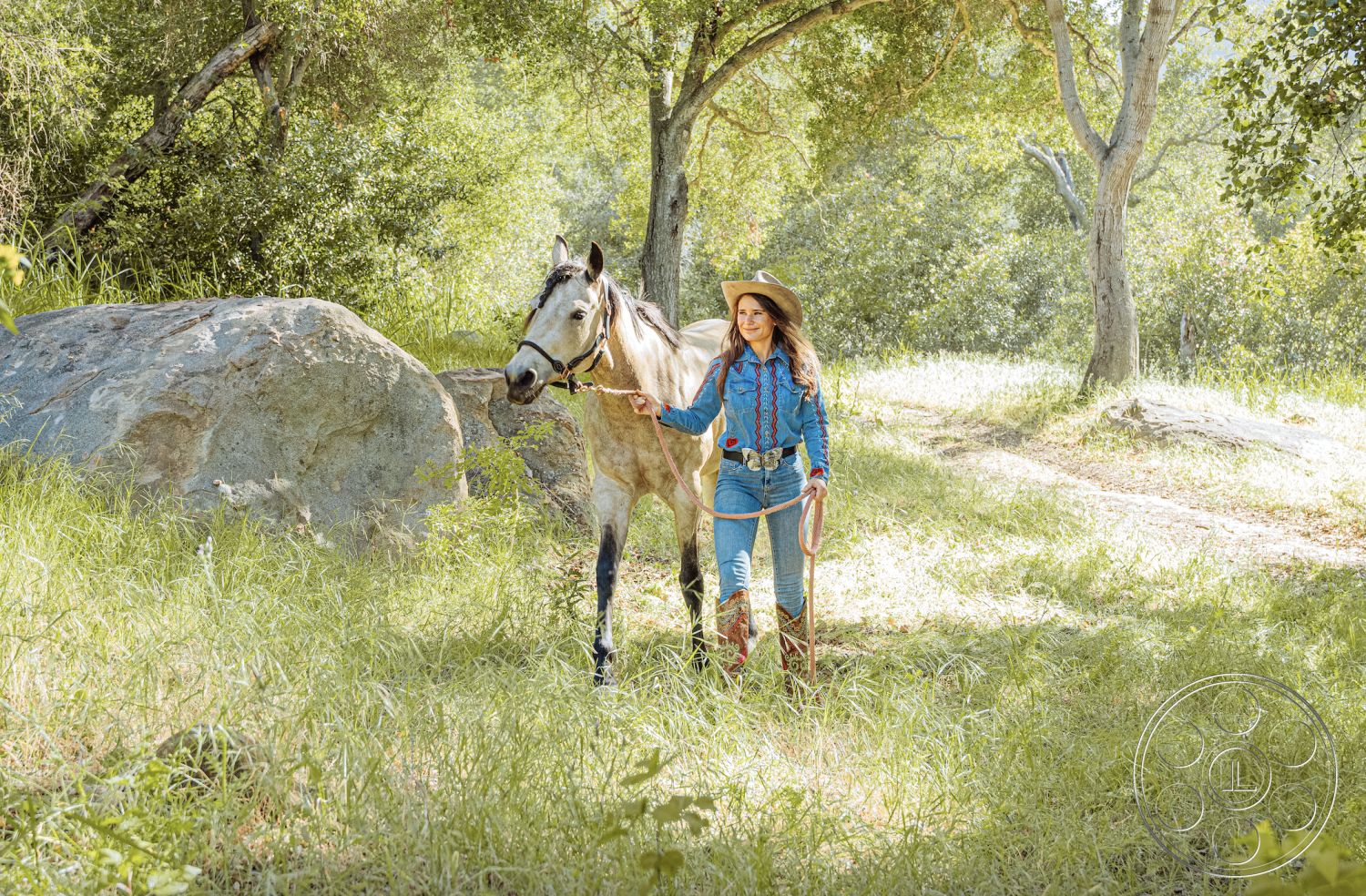 Topanga Retreat - sunlight filtering through trees, verdant foliage backdrop, natural grassland setting, outdoor rural environment, female equestrian in denim outfit, horse with a halter, weathered rock formation, cowboy boots and hat, horse riding path, rustic countryside atmosphere