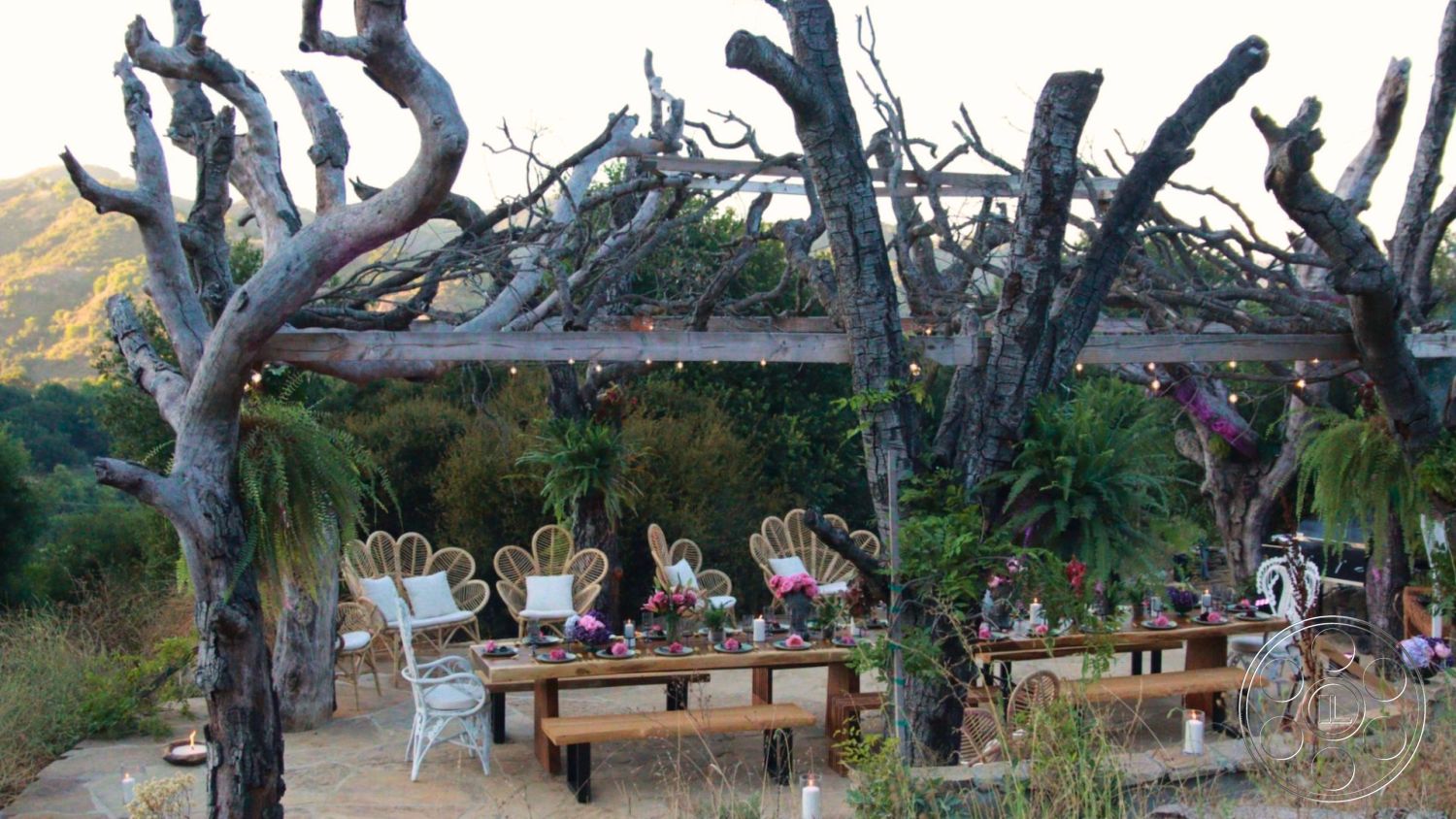 Topanga Retreat - natural materials, twinkling string lights, outdoor gathering space, open-air design, rustic outdoor dining area, landscape greenery, long wooden dining table, mountain view setting, natural stone patio flooring, woven rattan chairs, exterior wooden pergola, weathered wood beams, tropical ferns, decorative candle accents, casual entertaining area