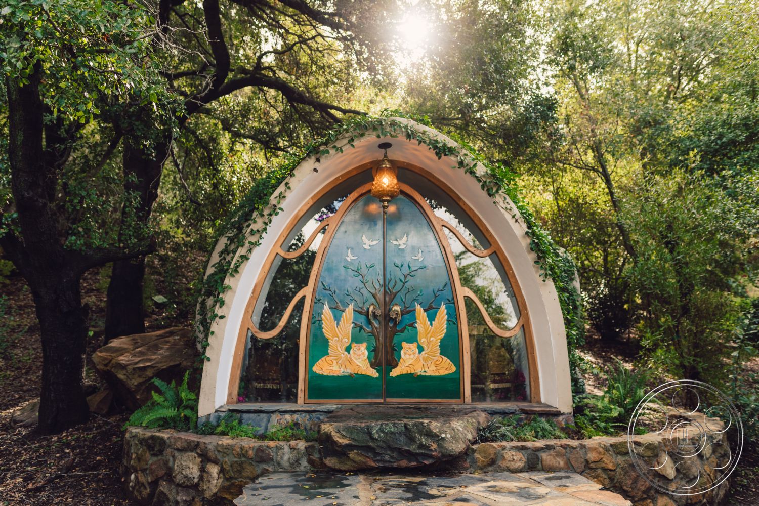 Topanga Retreat - exterior, surrounding greenery, natural stone wall, vine-covered walls, environmentally integrated design, flagstone pathway, decorative wooden door, arched entry door, colorful nature-inspired artwork, landscaped forest setting, rustic outdoor lighting fixture, organic architectural shape, multicolored paint accents, rounded structure, flowering plants alongside entrance