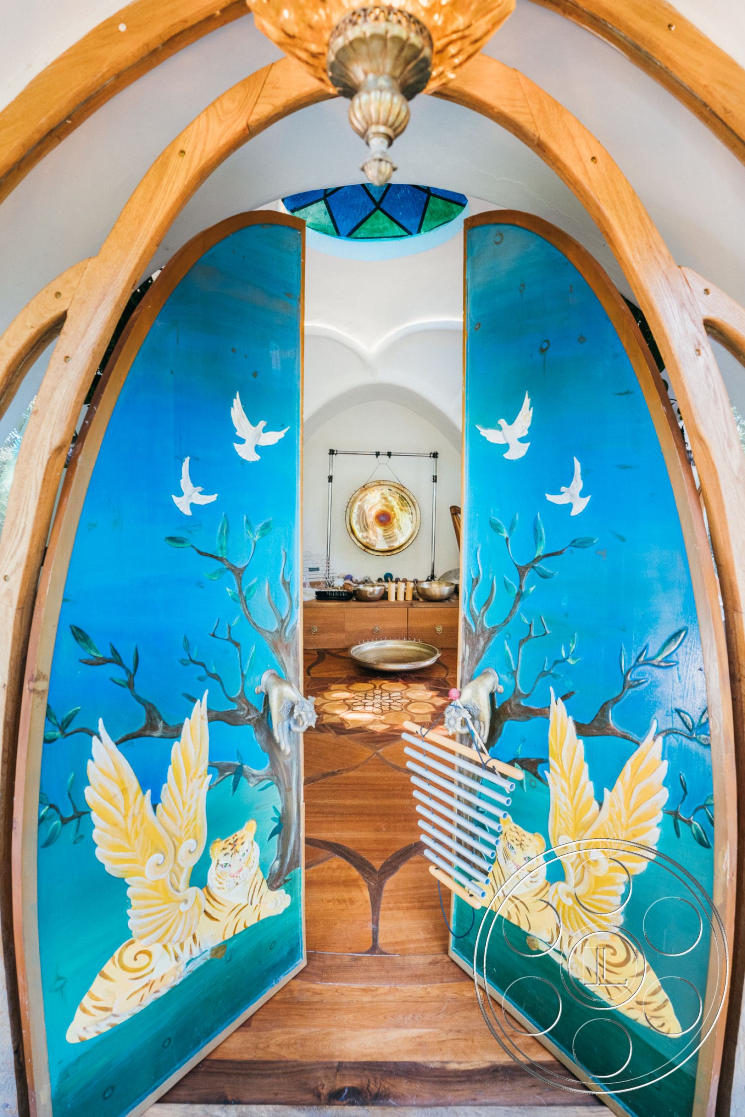 Topanga Retreat - open floor plan, natural wood flooring, inviting entrance, nature-inspired decor, handcrafted wood details, biophilic design elements, wooden arched front doors with colorful painted murals, light wood framing, interior with vaulted ceiling, ornate chandelier lighting fixture, decorative stained glass transom window, rustic kitchen setup, stone accents in the kitchen, wooden accent walls with artistic designs, flowing organic shapes through architectural design, bright and airy interior space, warm earthy color palette