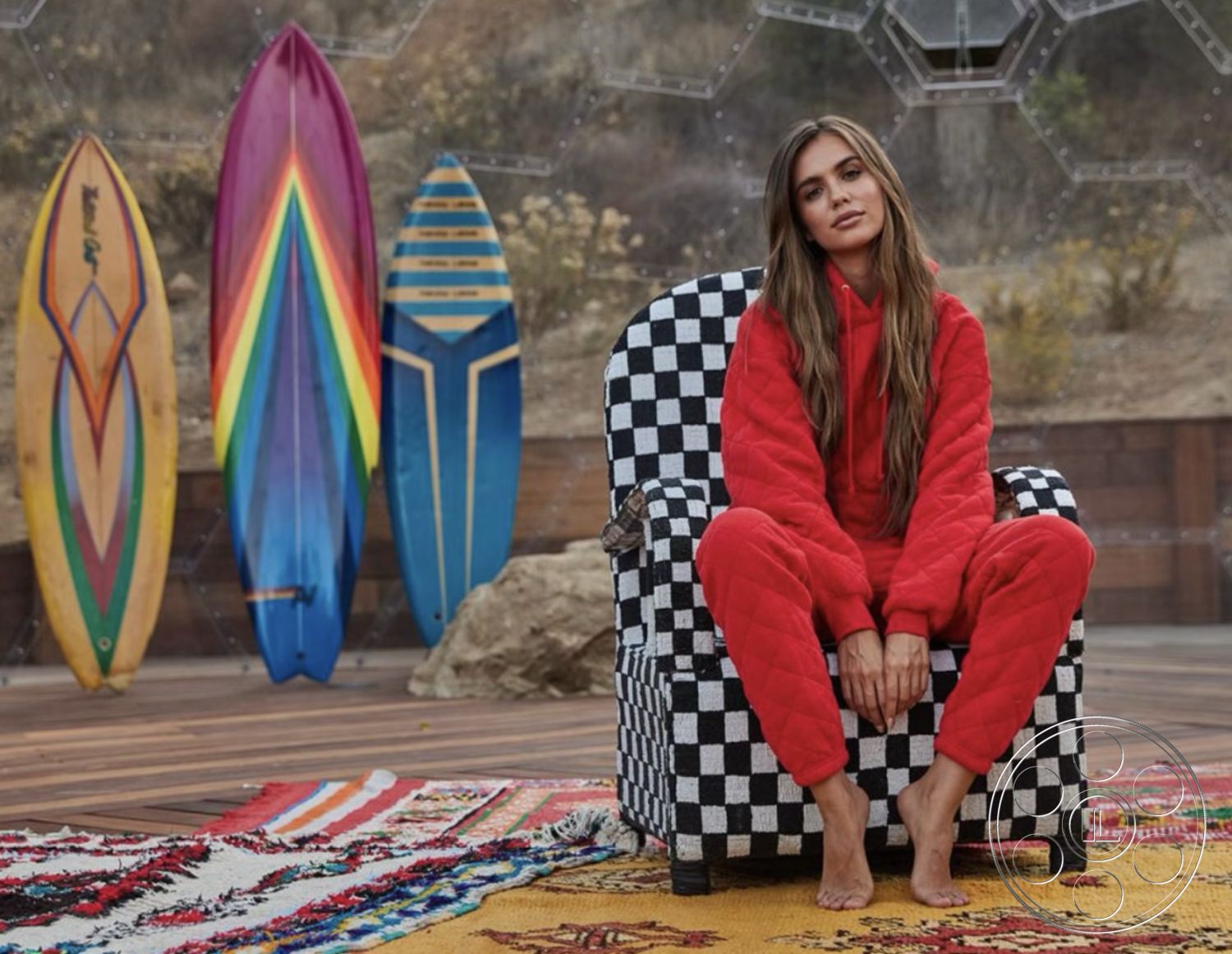 Topanga Retreat - outdoor seating area, bohemian aesthetic, mountain view setting, wooden deck flooring, sunlit outdoor space, open air environment, natural landscape backdrop, checkerboard patterned armchair, vibrant area rug, colorful surfboards, relaxed outdoor lifestyle, casual fashion style, red cozy outfit, modern eclectic decor, textured fabric chair