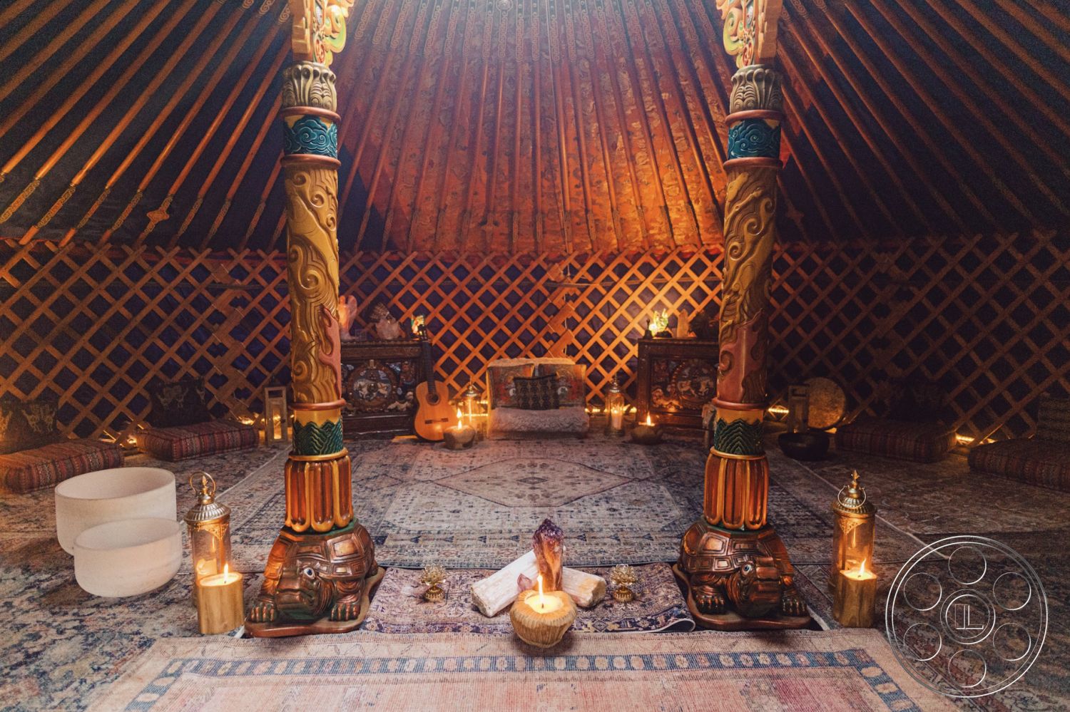 Topanga Retreat - intimate gathering space, meditative atmosphere, natural wood ceiling beams, interior yurt space, ambient candle lighting, wooden structural columns, hand-painted column details, woven lattice walls, colorful textile seating cushions, persian-style area rugs, yurt-style dome roof, earth-toned decor accents, acoustic guitar as decor, crystal bowls for sound therapy, decorative lanterns for illumination, traditional Mongolian design elements, sitting area with floor cushions, low lighting for ambiance, decorative wooden treasures