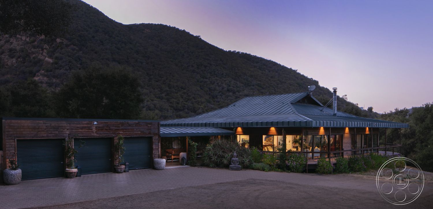 Topanga Retreat - open floor plan, rustic charm, concrete driveway, large floor-to-ceiling windows, outdoor lighting fixtures, environmentally integrated design, mountain backdrop setting, landscaped garden patio, modern architectural design, metal roof panels, exterior wooden siding, wraparound deck, earth-toned stone pavers, natural stone landscape accents, three-car garage with roll-up doors, bushes and drought-resistant plants