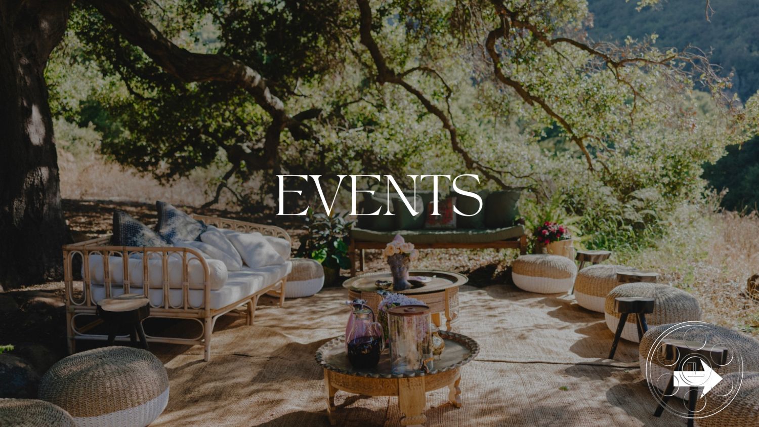 Topanga Retreat - outdoor seating area, wooden side tables, white upholstered sofa, relaxed outdoor ambiance, botanical setting, woven poufs, earth-toned decor, natural wood coffee table, lush greenery background, tan jute rug, rustic event space, sun-dappled seating arrangement, garden party atmosphere, organic shapes in furnishings, casual outdoor lounge, flower arrangements in glass vases, comfortable event seating, semi-covered area under trees, scenic natural backdrop, event setting in the woods