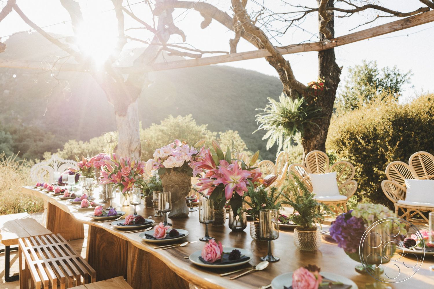 Topanga Retreat - wooden dining table, outdoor seating area, natural landscape view, wildflower garden, woven rattan chairs, wooden trellis structure, exterior patio setting, rustic wooden benches, patterned ceramic plates, glass vases with flowers, overhead string lights, textured wooden beams, sunlit outdoor dining, large mountain backdrop, decorative centerpiece arrangements, seasonal floral decorations, natural wood surface