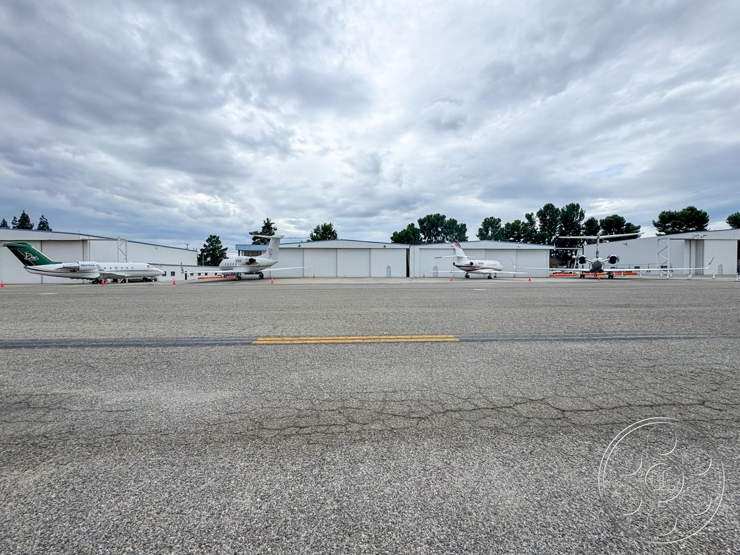 VN Hangar - large hangar doors, asphalt runway surface, metal hangar structures, flat-roof hangar buildings, concrete aircraft parking area, aircraft tarmac, cloudy sky setting, aviation facilities exterior, commercial jet and private aircraft, single-story hangar design, exposed metal framework, airfield environment, landscaped periphery with trees