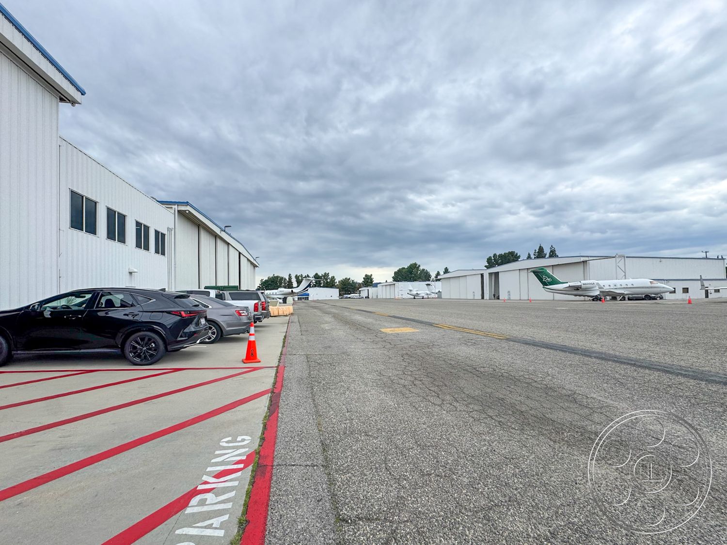 VN Hangar - cloudy sky backdrop, concrete parking lot surface, exterior airport hangar with metal siding, asphalt runway markings, rectangular airplane parking spaces, striped red parking lines, flat-roofed aircraft hangar, large glass windows on hangars, landscape of surrounding trees, fenced aircraft area, vehicles in parking lot, modern corporate jets on tarmac, open space adjacent to hangars