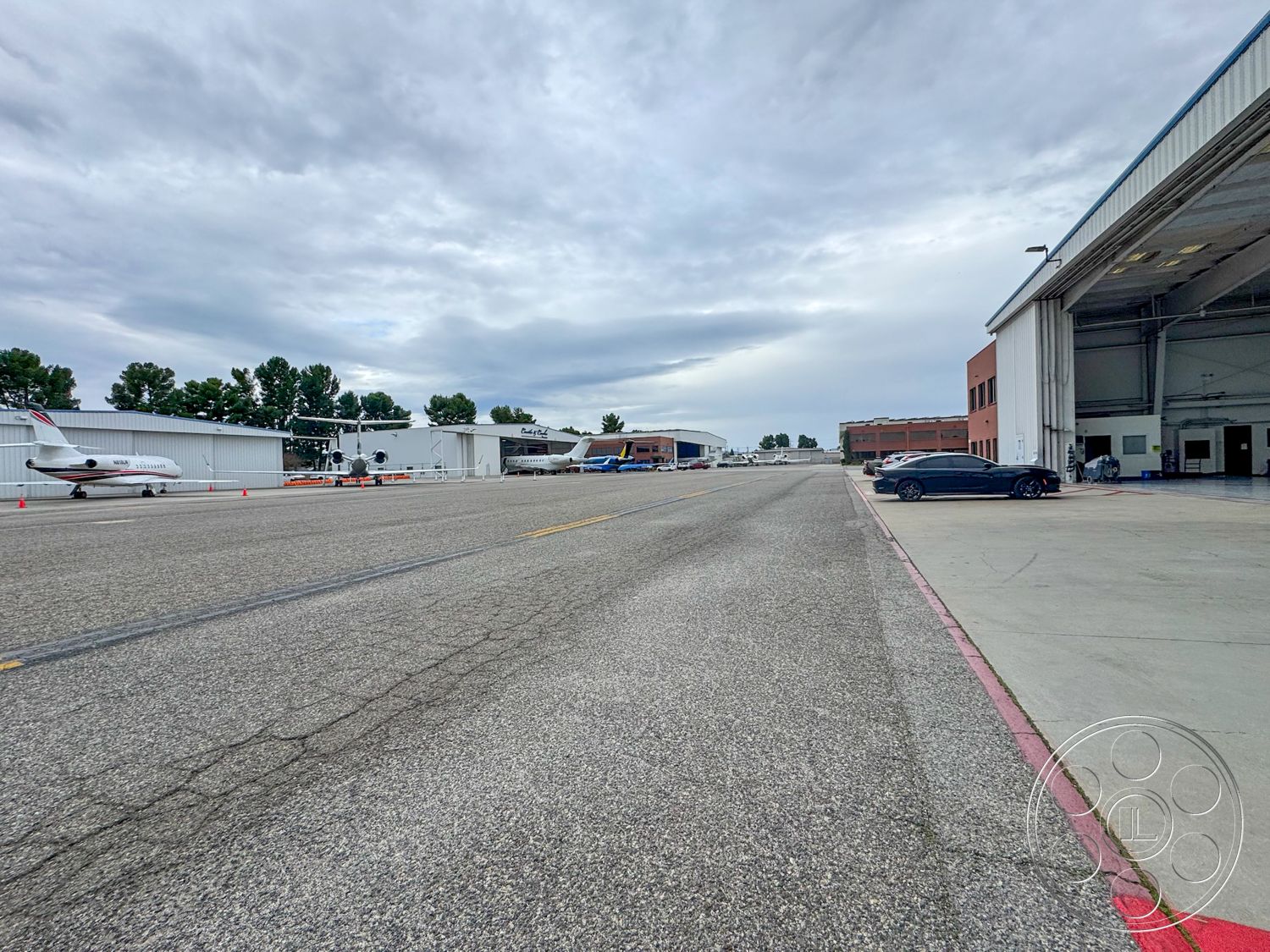 VN Hangar - paved parking lot, flat roof design, cloudy sky backdrop, surrounding tree line, urban airport setting, asphalt runway surface, concrete taxiway apron, hangar metal siding, single-story aviation hangar, industrial steel structure, high-clearance bay doors, exterior loading area, curved drive access, fenced airport perimeter, multiple small aircraft parking, drainage grates in pavement