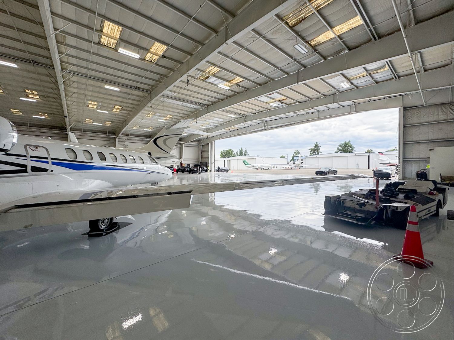 VN Hangar - industrial lighting fixtures, concrete slab flooring, interior open space layout, exterior hangar structure, aircraft parking area, metallic steel frame, high ceiling with exposed trusses, glass-paneled hangar doors, reflective epoxy floor finish, maintenance equipment storage, large movable service tugs, safety cones for vehicle access, blend of natural and artificial light, airplane runway view, airport facility environment