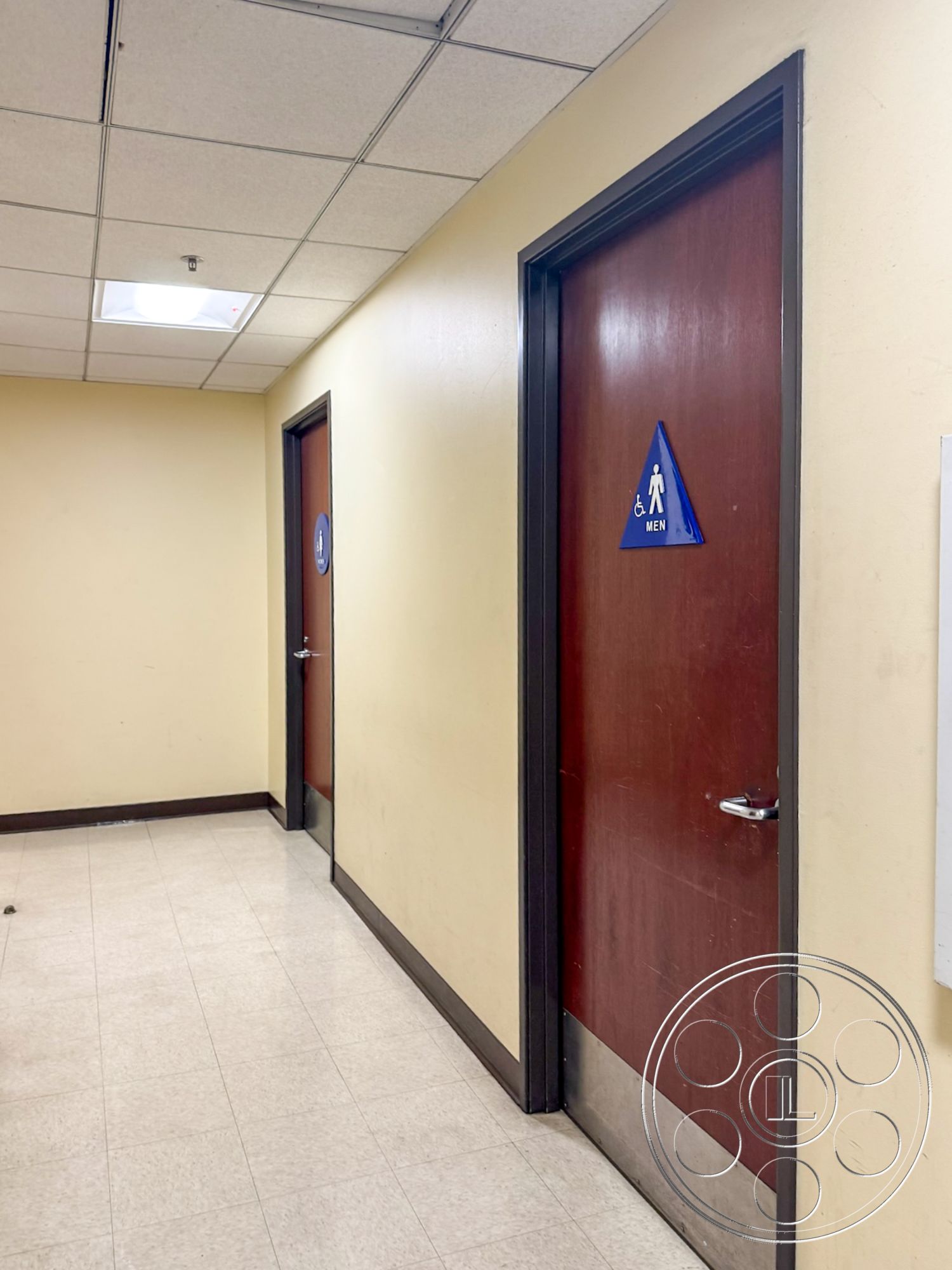 VN Hangar - brown wooden doors, interior hallway, dimly lit corridor, drop ceiling tiles, fluorescent ceiling lights, beige painted walls, metal door handles, vinyl floor tiles, non-slip flooring, ADA compliant signage, public restroom entrance, accessible restroom