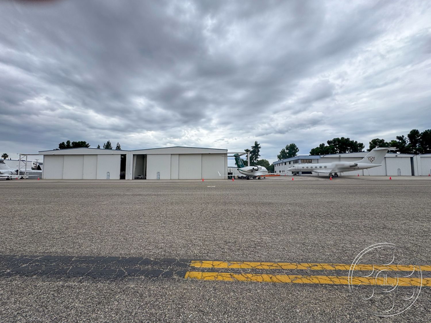 VN Hangar - surrounding tree line, asphalt runway surface, exterior metal hangar building, white corrugated metal walls, commercial aircraft parking area, flat-roofed hangar design, open aircraft storage space, landscaped airport grounds, overcast sky environment, ground-level view of transport aircraft, sliding hangar doors, aircraft taxiway