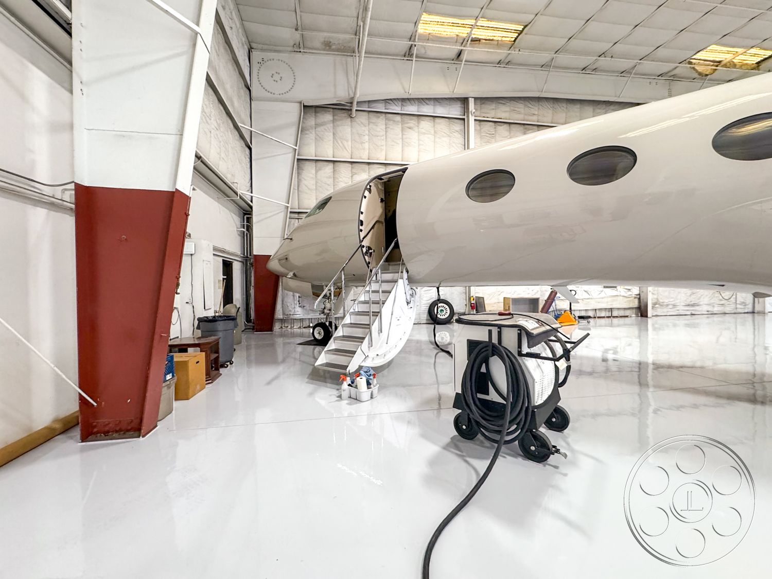 VN Hangar - industrial storage space, large glass windows for natural light, aircraft maintenance area, metal framed structure interior, white epoxy-coated hangar floor, large aircraft access door, exposed steel beam construction, hangar with high vaulted ceiling, conventional workshop area, arrangement of tools and equipment, access stairs to aircraft interior, utility cart with cleaning supplies, well-lit aircraft hangar, large enclosed space for aircraft, functional and practical design