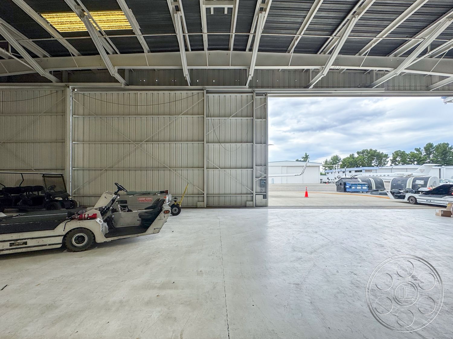 VN Hangar - concrete flooring, large windows providing natural light, spacious work environment, high ceilings with exposed trusses, exterior steel warehouse structure, metal roofing panels, open bay door design, industrial space layout, mechanized warehouse equipment, utility vehicle parking area, outdoor storage area visible, surrounding green tree line, clear sky background