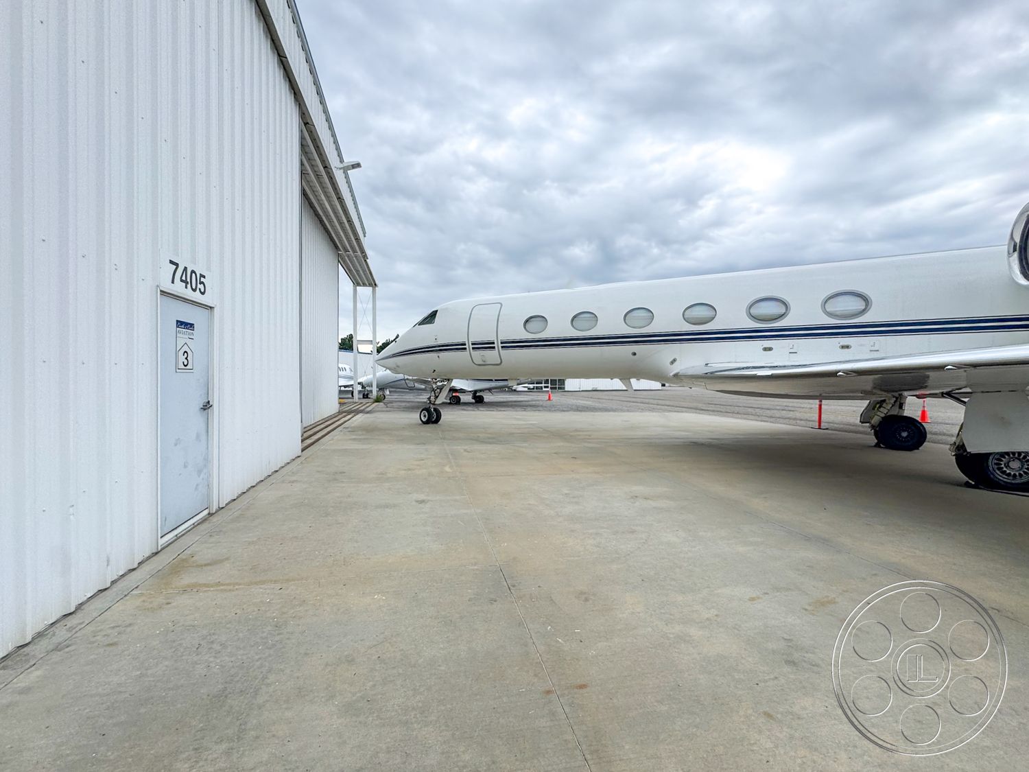 VN Hangar - commercial aviation facility, exterior steel airplane hangar, white corrugated metal siding, concrete taxiway, single airplane parking space, large private jet, gray overcast sky, modern aircraft design, door number 7405, industrial aviation environment, expansive airfield view