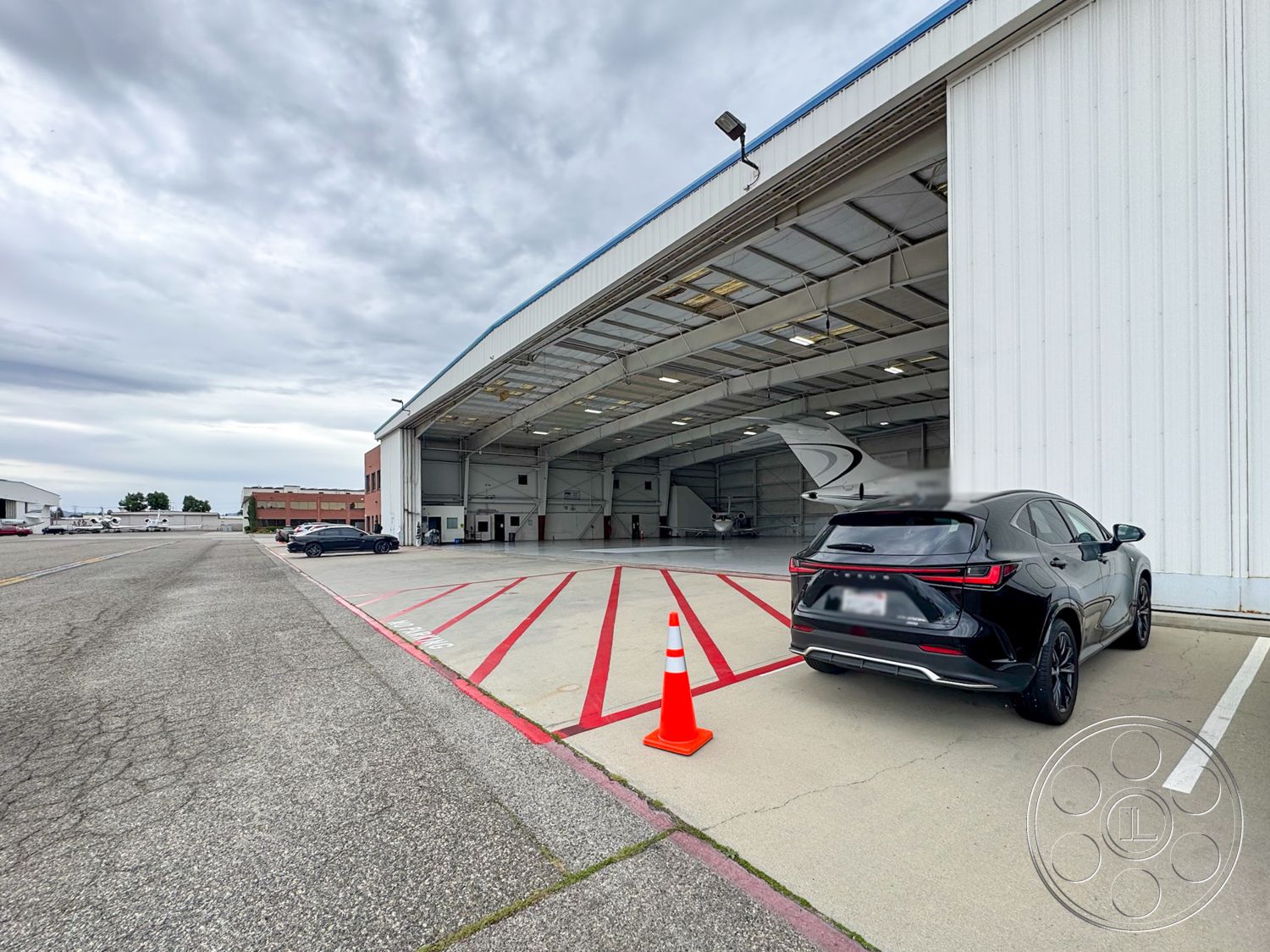 VN Hangar - large open space, exposed metal beams, concrete parking lot, automobile parking area, metal hangar exterior, sliding hangar doors, steel framed hangar structure, asphalt access road, striped parking markings, industrial aircraft storage, gray sky backdrop, urban airport environment, flat-roof commercial building, operational space for aircraft, nearby aircraft tarmac