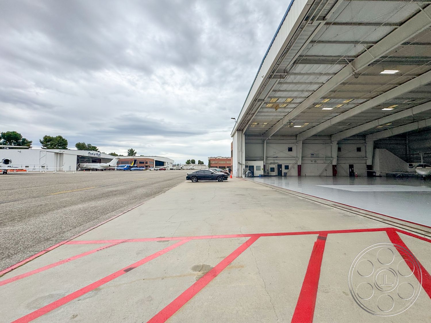 VN Hangar - exterior, mountainous backdrop, asphalt runway, cloudy sky, gray concrete surface, aircraft parking area, commercial aviation facility, large overhead lighting fixtures, concrete apron, steel hangar structure, large industrial hangar doors, glazed windows in hangar, painted red safety lines, modern aircraft hangar design, surrounding airport setting, aircraft service area, concrete loading area, industrial drainage strips, paved taxiway access