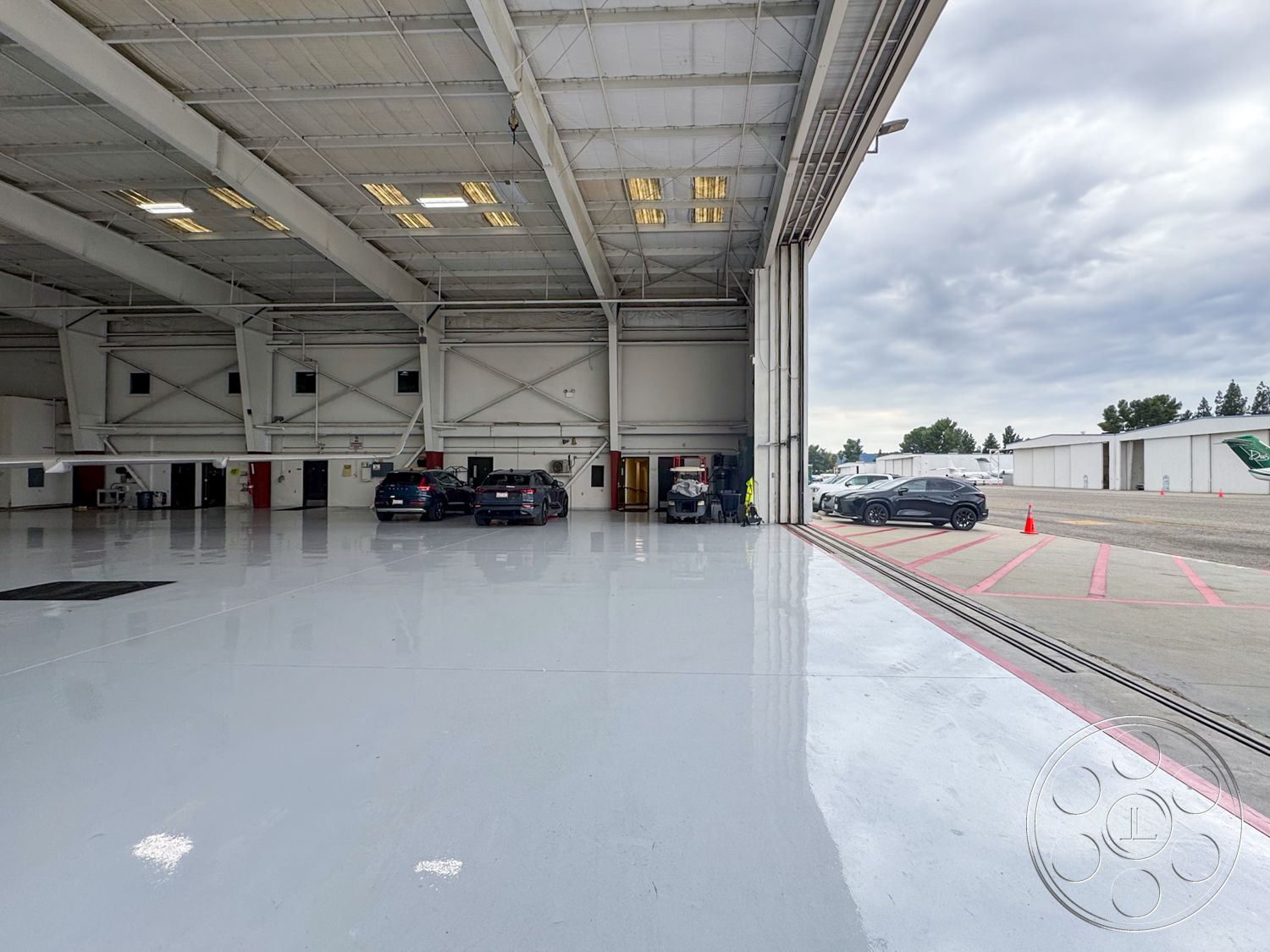 VN Hangar - industrial lighting fixtures, high vaulted ceiling, maintenance area, ground-level access, white painted walls, steel frame construction, exterior hangar structure, commercial aviation facility, direct access to runway, large roll-up doors, open hangar space, epoxy coated concrete flooring, metal cladding exterior, automotive parking section, surrounding airport environment, gray skies setting, concrete apron area, vehicle storage area
