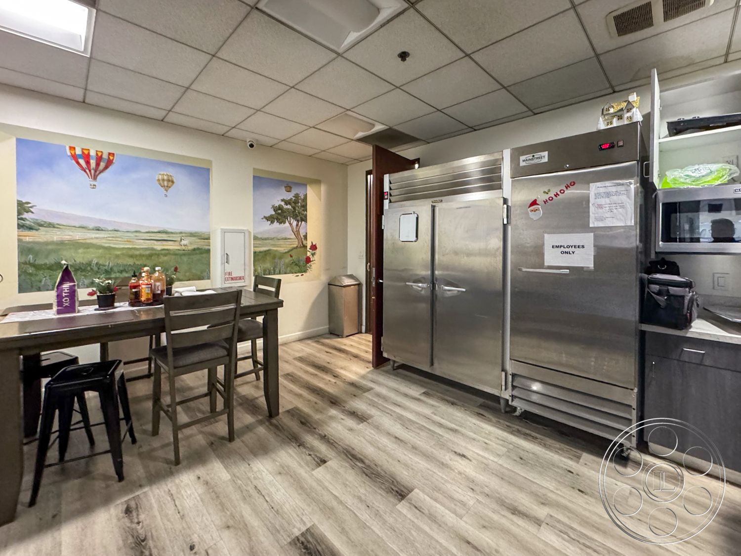 VN Hangar - potted plant decor, interior kitchen space, open kitchen layout, stainless steel microwave, metal trash can, stainless steel commercial refrigerator, wood dining table with high stools, laminate floorboards, lightly textured ceiling tiles, wall-mounted artwork featuring hot air balloons, white painted walls with mural, dark gray cabinet doors, functional employee break room