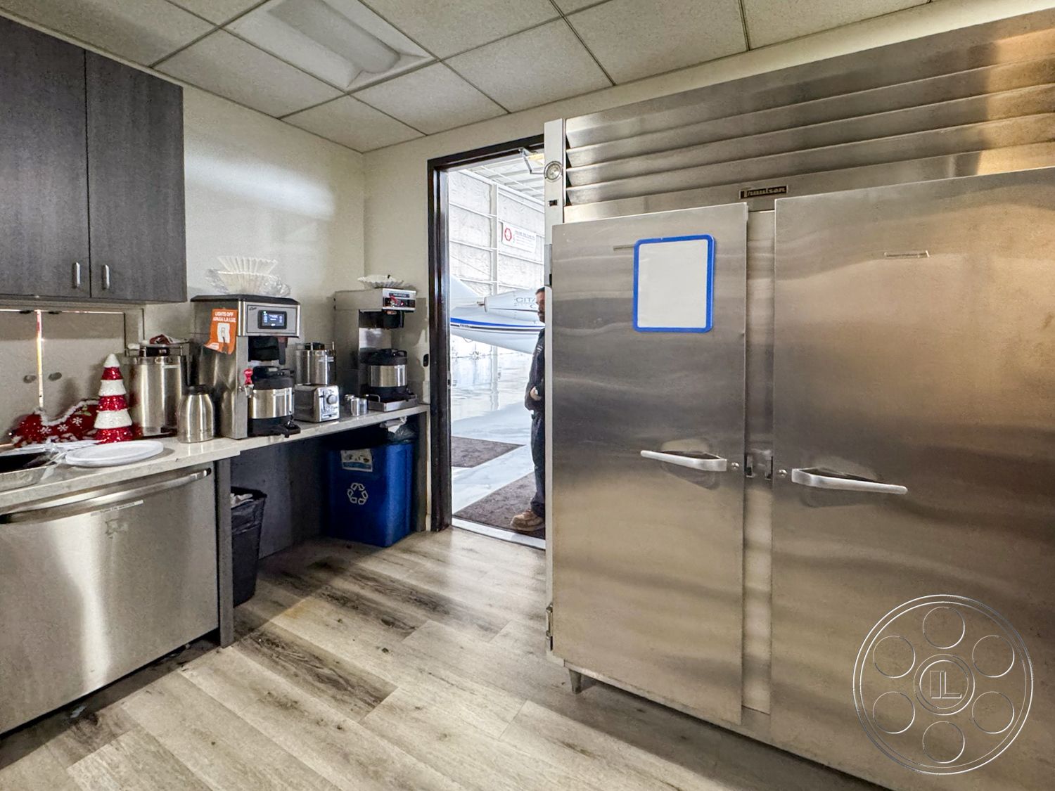 VN Hangar - interior kitchen space, open kitchen layout, stainless steel refrigerator doors, laminate countertop surface, wood vinyl flooring, modern cabinetry with dark finish, utility area with appliances, commercial coffee maker station, kitchen entry with glass door, decorative holiday elements, recycling bin for waste management, multiple beverage dispenser units, bright overhead ceiling lights