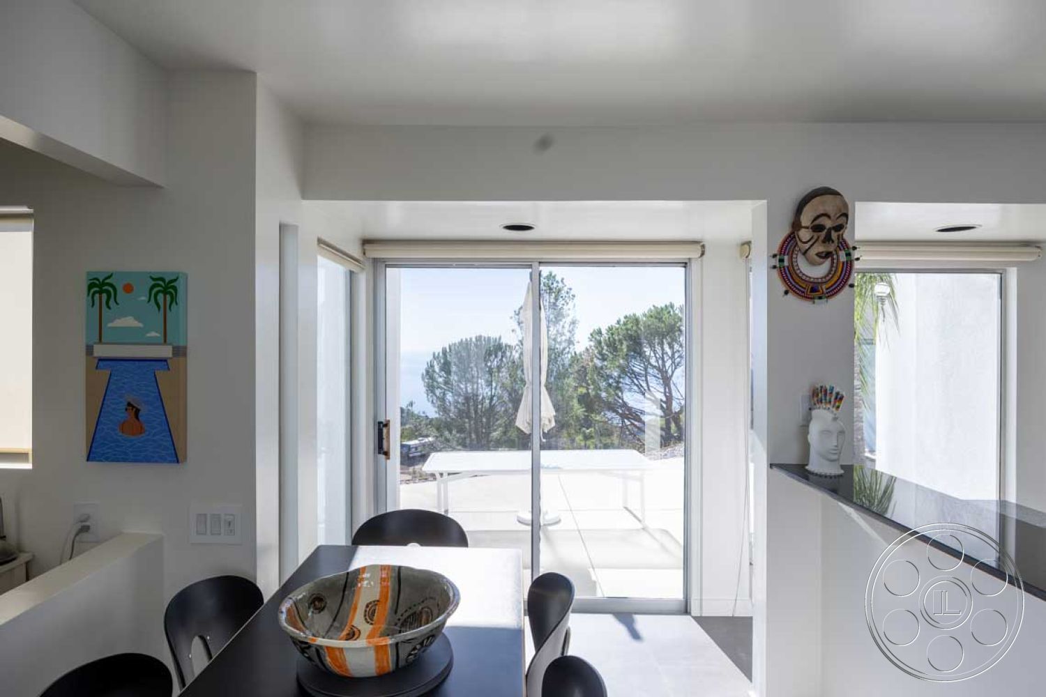 Modern 56 - stainless steel kitchen appliances, black granite kitchen countertop, open floor plan living area, floor-to-ceiling windows with ocean view, interior kitchen with white painted walls, glass sliding doors leading to outdoor space, exterior patio with concrete flooring, white ceiling with recessed lighting, contemporary artwork on wall, modern dining room with circular table, minimalist decor with natural light, wooden hanging light fixture, black round dining chairs, simple bowl centerpiece on dining table, modern coastal setting with surrounding trees, light and airy atmosphere with neutral tones