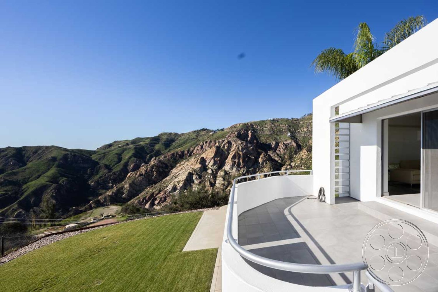 Modern 56 - white stucco walls, integrated landscaping, flat roof design, mountain view setting, open-concept living area, contemporary outdoor living space, floor-to-ceiling glass sliding doors, modern minimalist architecture, exterior deck with concrete flooring, curved balcony with glass railings, landscape with green grass, sustainable hillside location, smooth concrete patio surface