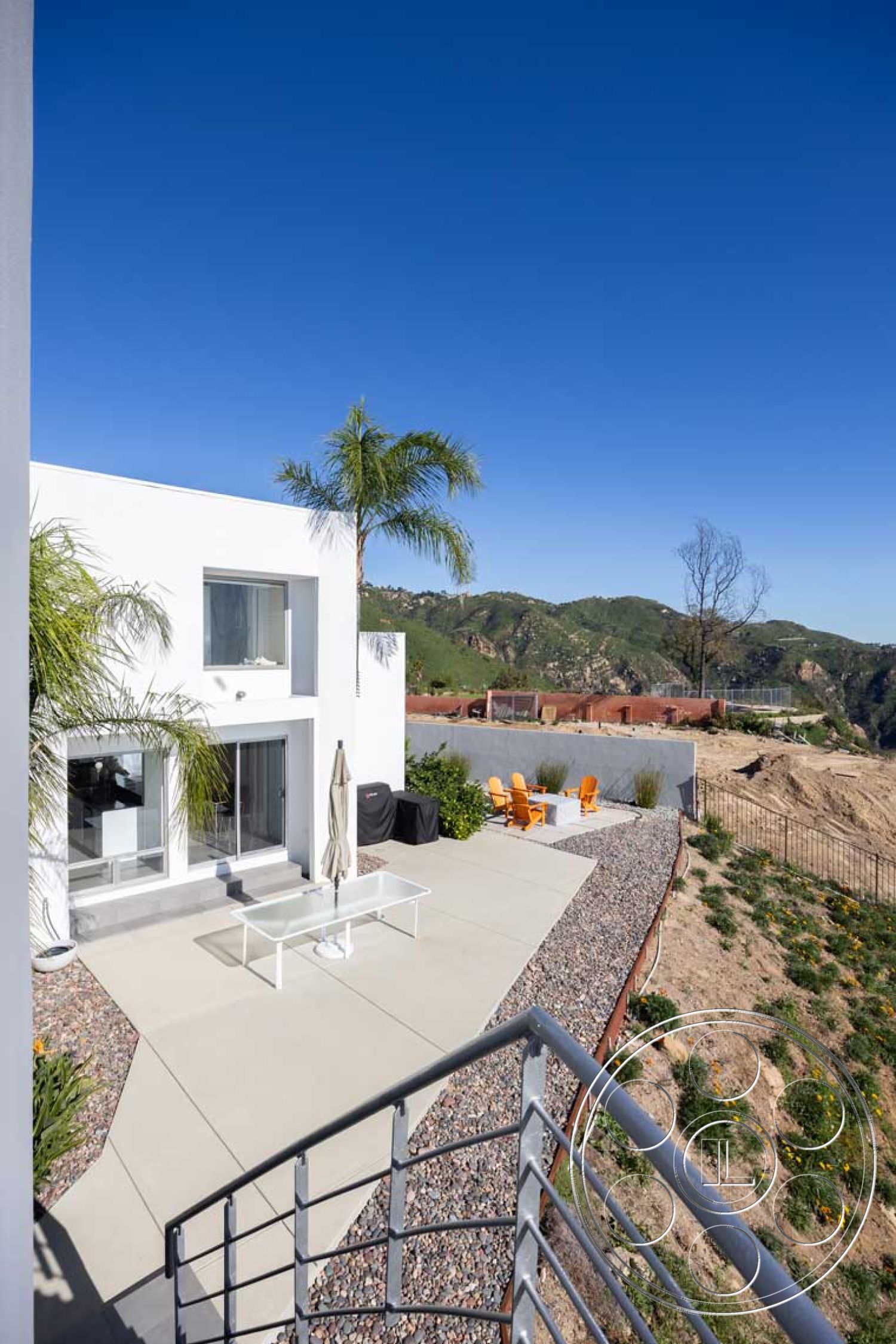 Modern 56 - exterior, white stucco walls, glass sliding doors, contemporary outdoor furniture, sunny outdoor area, mountain view setting, concrete patio flooring, gravel border around garden, modern minimalist architecture, palm trees landscaping, steel railing details, open-plan design, rectangular patio, sloped hillside, exposed aggregate border, landscaped garden with boulders