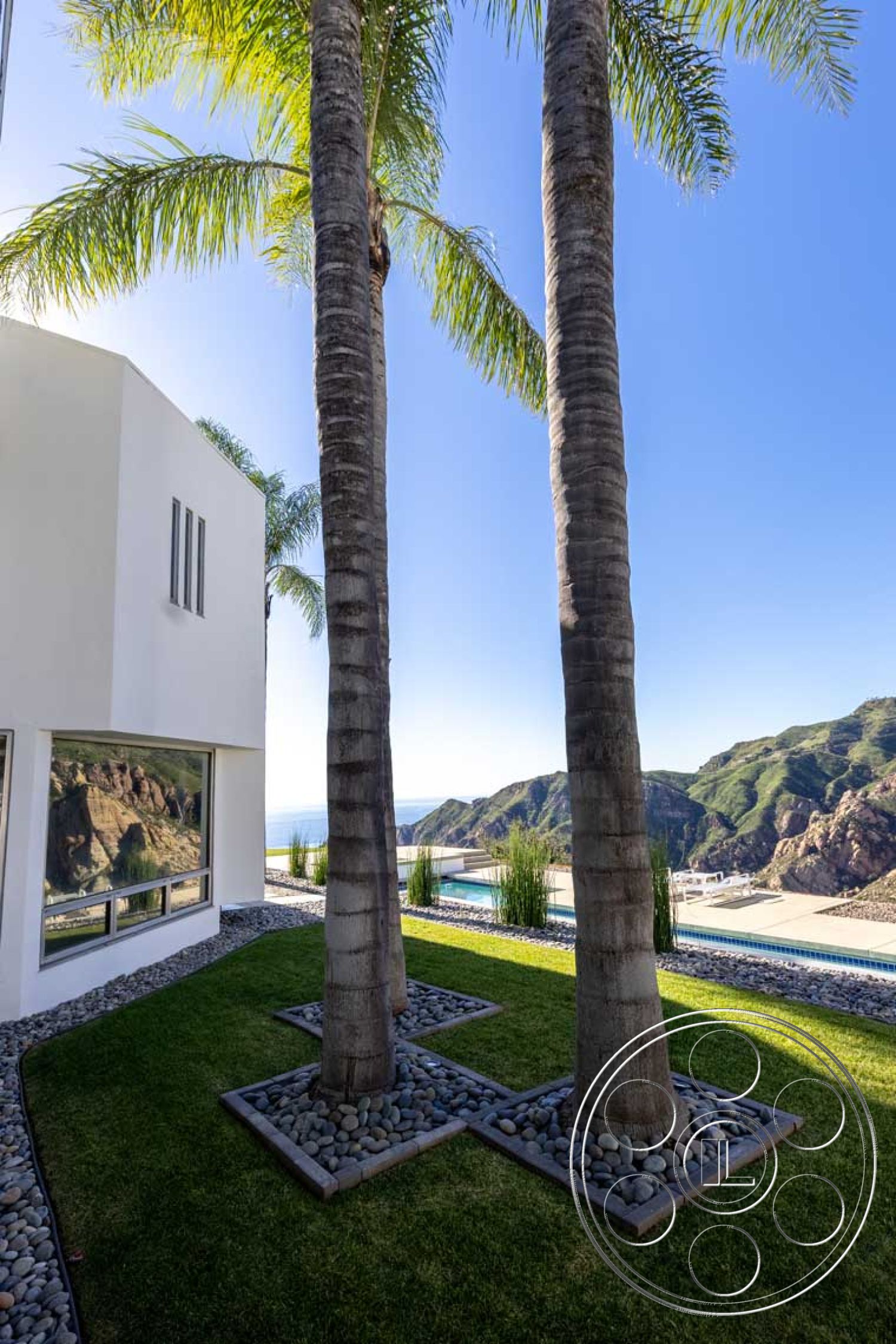 Modern 56 - exterior, modern architecture, minimalist design, large glass windows, concrete wall, outdoor living space, landscaped lawn, mountain backdrop, hillside location, gravel ground cover, tree-lined property, palm trees landscaping, stone garden border, pool area with ocean view, slim rectangular pool, sleek white facade