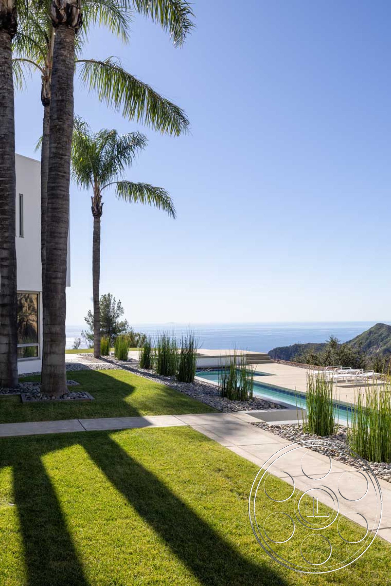 Modern 56 - exterior, ocean view, concrete walkway, open sky, sloping hillside, sunny outdoor setting, modern minimalist design, concrete patio floor, rectangular infinity pool, landscaped garden with green grass, palm trees in foreground, natural stone border around pool, surrounding coastal environment, resort-style outdoor area