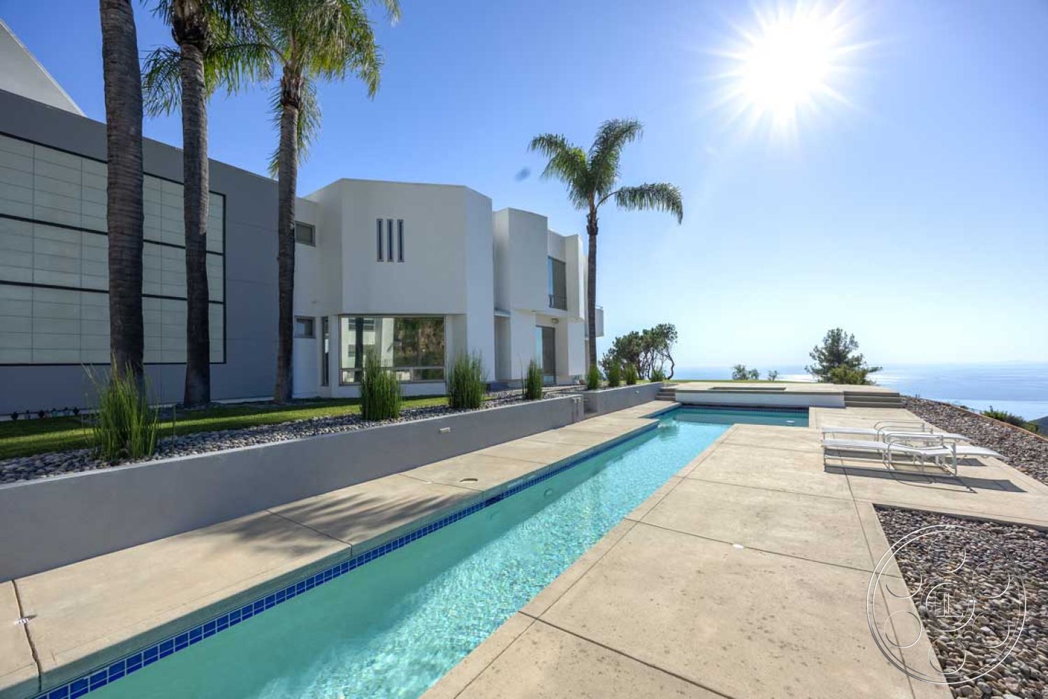 Modern 56 - exterior, horizontal siding, glass sliding doors, floor-to-ceiling windows, outdoor lounge area, modern architectural style, gravel landscaping, concrete pool deck, rectangular pool, smooth concrete walls, concrete patio floor, landscape with palm trees, infinity pool edge, minimalistic design features, oceanview setting, sunny cloudless sky, sloped yard
