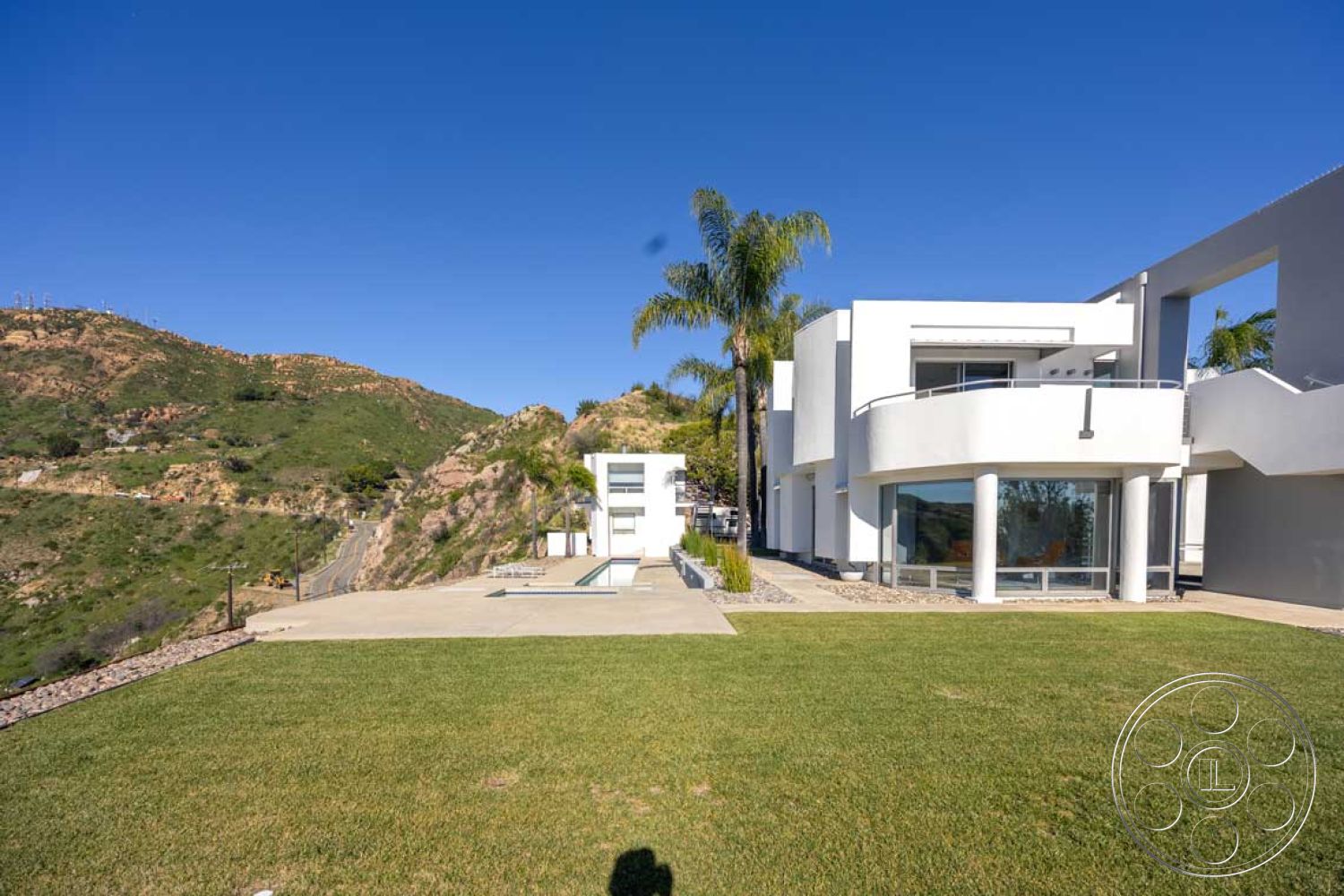 Modern 56 - minimalist design elements, modern architectural style, open outdoor space, smooth concrete driveway, exterior white stucco walls, floor-to-ceiling glass windows, infinity pool with concrete border, contemporary residential architecture, surrounding mountain view, sloping hillside location, rectangular concrete patio floor, landscaped green grass lawn, curved white balcony railings, palm trees in landscape design, stylish exterior entryway