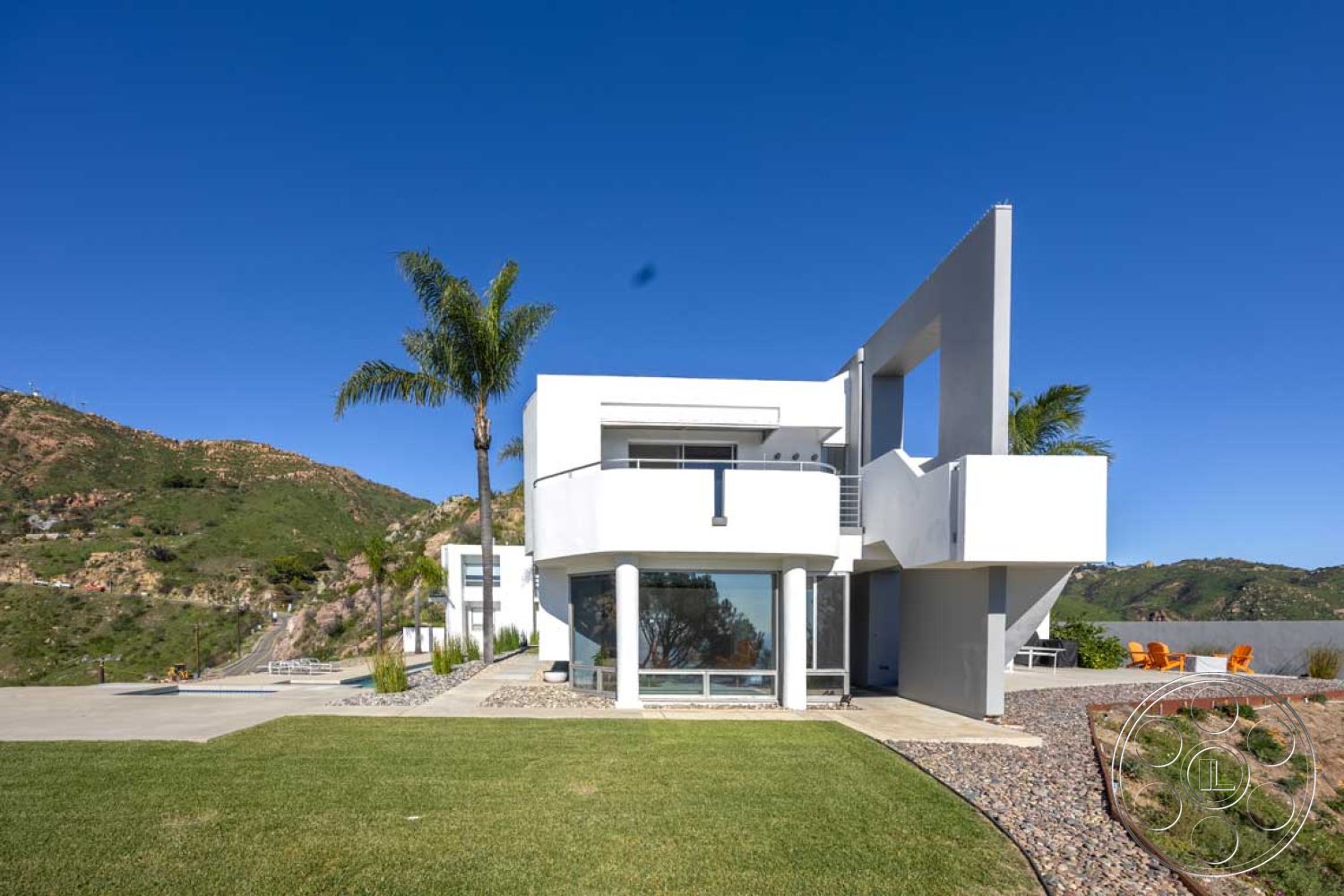 Modern 56 - exterior concrete driveway, interior open concept layout, interior large floor-to-ceiling windows, exterior stone pathway, exterior white stucco walls, interior minimalist design, interior polished concrete floors, exterior hillside view, exterior flat roof, exterior geometric modern architecture, exterior expansive glass windows, exterior landscaped garden with grass, exterior palm trees and shrubs, interior sleek modern finishes, exterior outdoor seating area with orange chairs, exterior pool area with unobstructed views