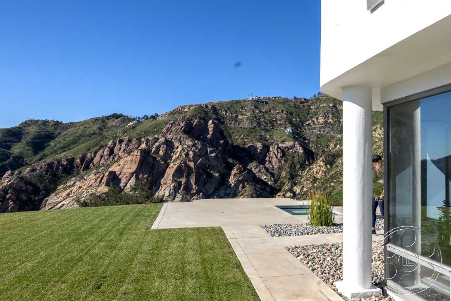 Modern 56 - exterior, white stucco walls, modern architecture, minimalist design, large glass doors, floor-to-ceiling windows, outdoor living space, concrete patio flooring, recessed lighting fixtures, contemporary home design, natural stone landscaping, open outdoor terrace, landscaped grass garden, sloped mountain backdrop, stone border detailing, infinity pool area, view of mountain range