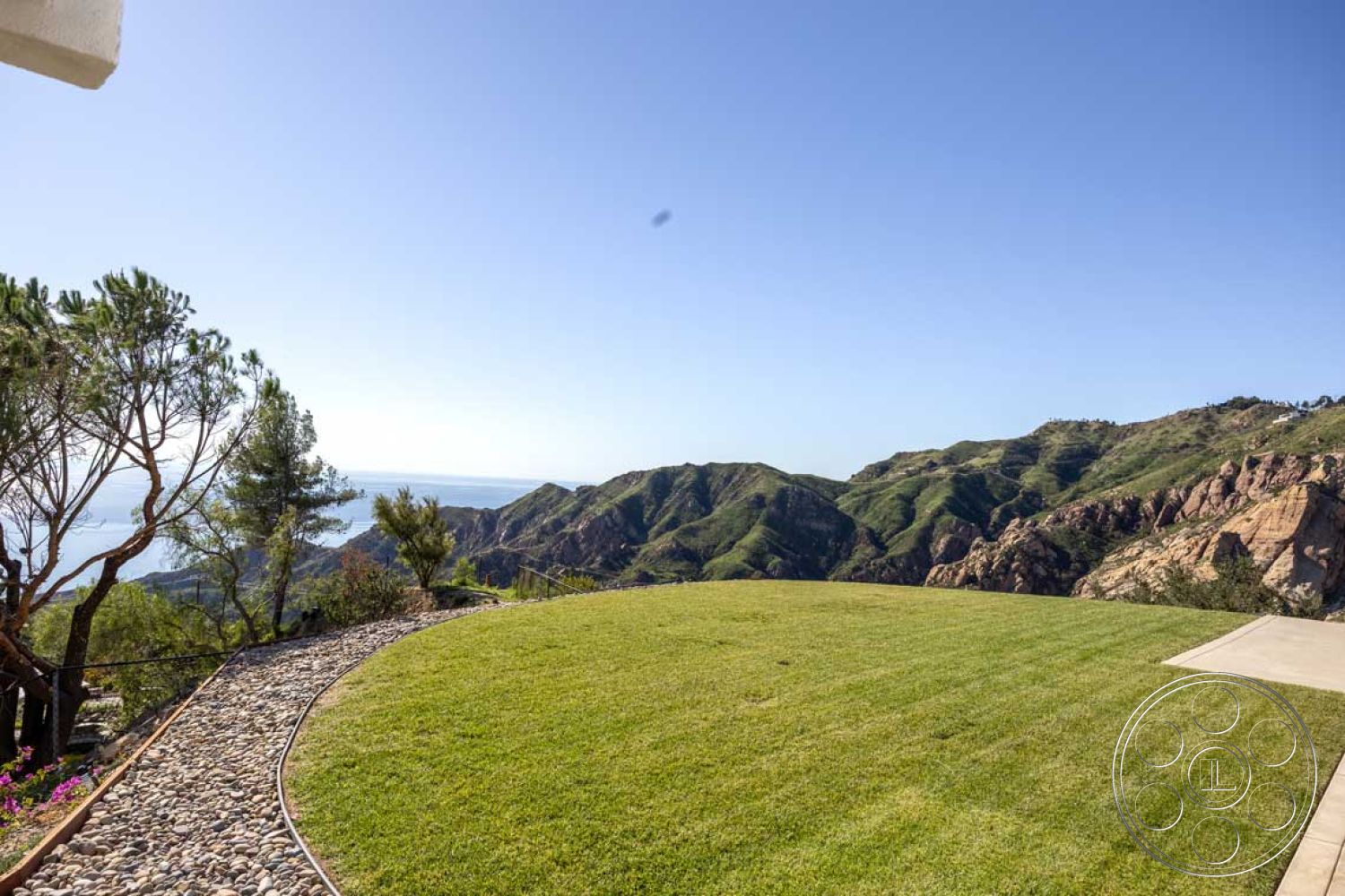 Modern 56 - outdoor living space, lush greenery surroundings, exterior lawn with manicured grass, gravel pathway with smooth pebbles, natural hillside landscape, ocean horizon view, mountainous terrain backdrop, curved patio edge, California coastal setting, sunny clear sky