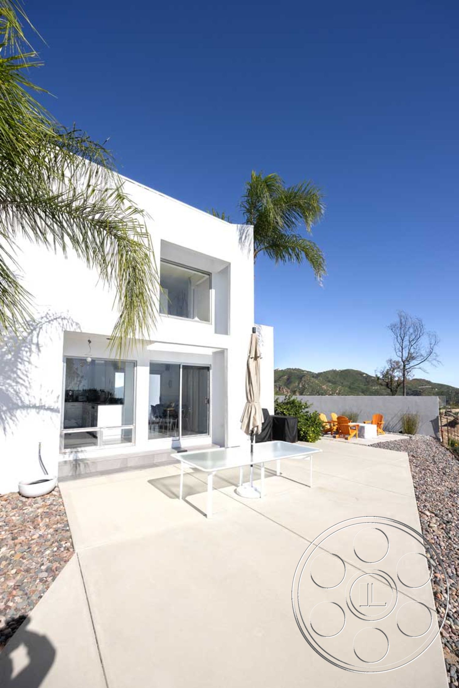 Modern 56 - exterior, natural stone accents, white stucco walls, outdoor dining area, modern architectural style, sunshade umbrella, mountain view setting, floor-to-ceiling sliding glass doors, concrete patio floor, minimalist outdoor furniture, palm tree landscaping, rectangular patio, clear blue sky backdrop, landscaped gravel border