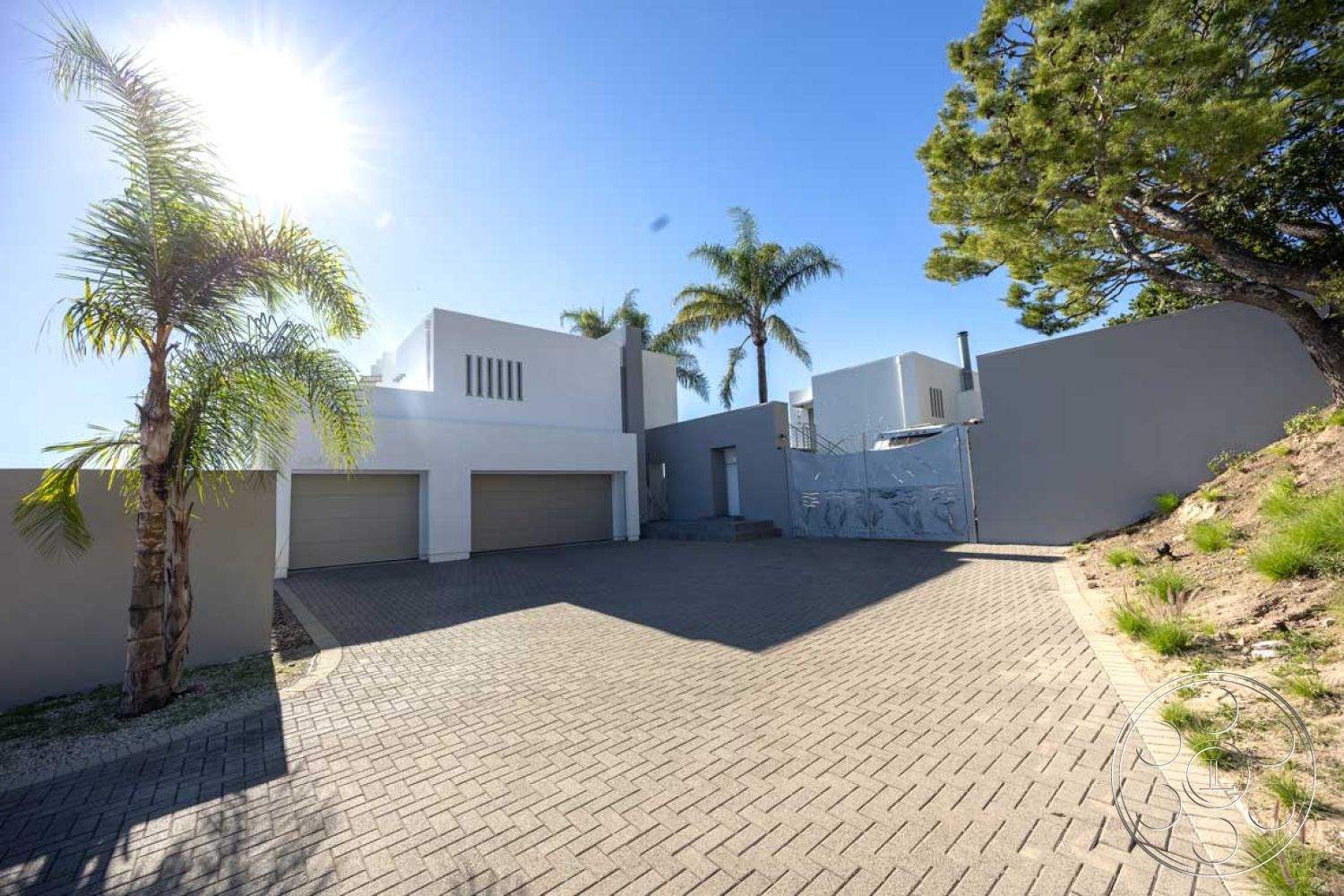 Modern 56 - exterior, gray color scheme, sunlit facade, minimalist architectural style, surrounding hillside vegetation, tropical landscaping with palm trees, smooth stucco walls, flat roofline, paved driveway with interlocking brick pavers, contemporary garage doors, modern entry gate with artistic design, curved driveway approach, nearby residential setting