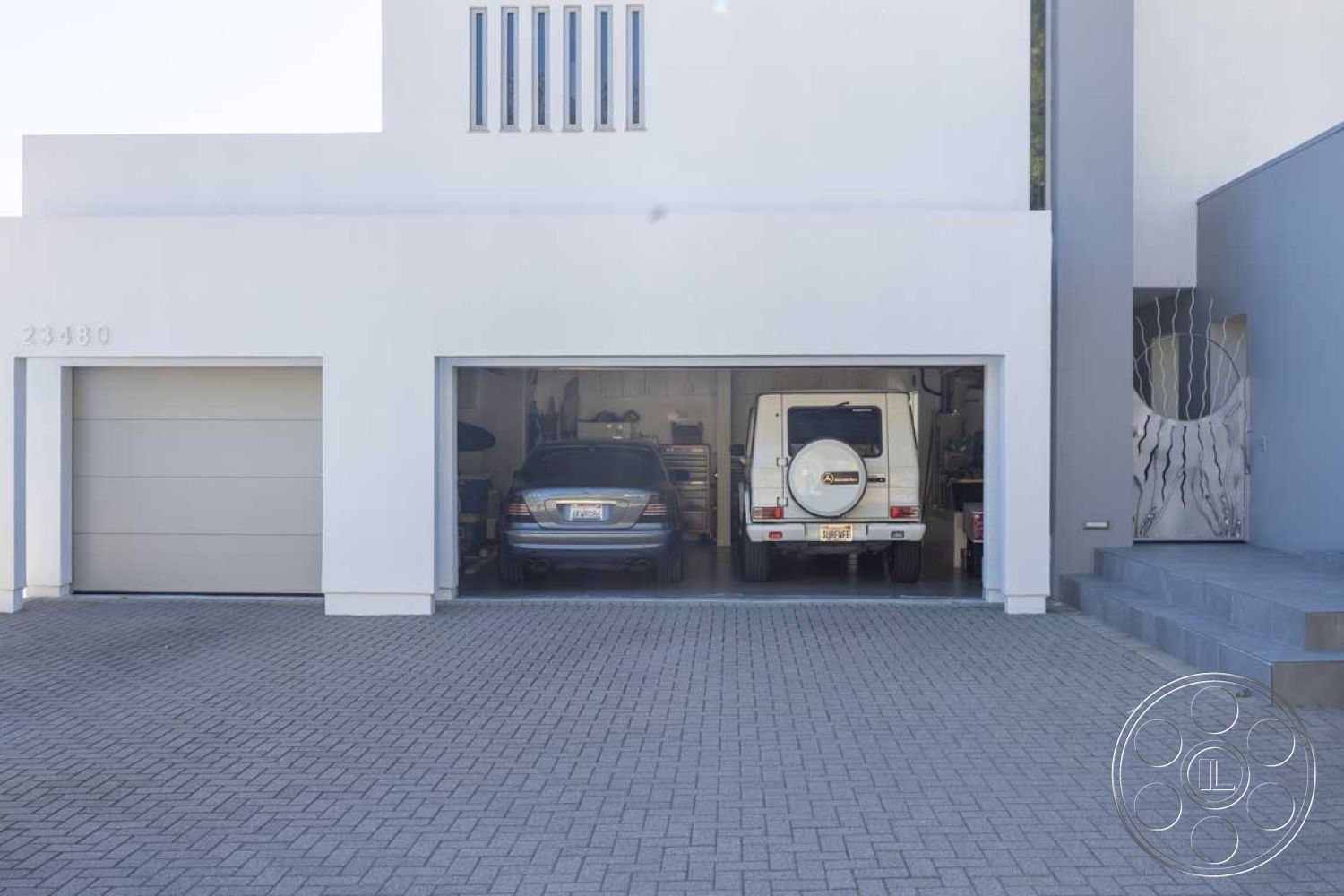 Modern 56 - low-maintenance landscaping, concrete driveway, two-car garage, geometric shapes in architecture, interior garage space, exterior light fixtures, exposed framing elements, urban suburban setting, curved driveway entrance, smooth stucco exterior walls, modern exterior finishes, modern minimalist architectural style, paved herringbone driveway, gray metal garage doors, asymmetrical home design, integrated outdoor steps, painted house number, glass accent panels, horizontal window configurations, concrete block retaining wall, open garage facing exterior