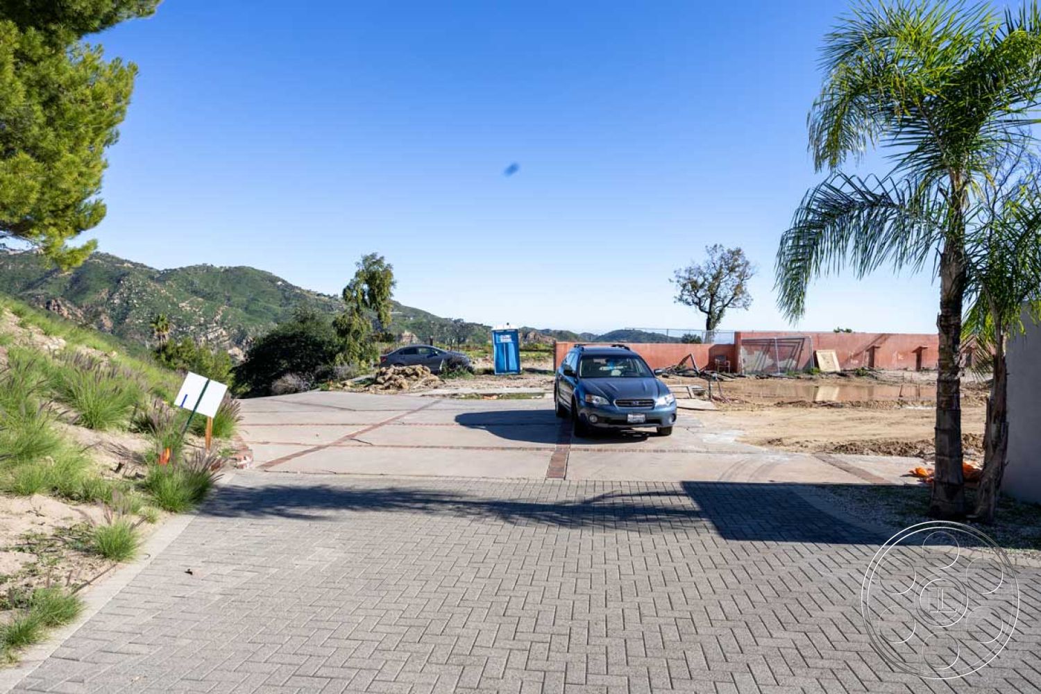 Modern 56 - clear blue sky, green grass landscaping, surrounding mountain view, interior concrete driveway, paver driveway stones, exterior suburban landscape, decorative brick edging, bordering palm trees, construction site backdrop, partial hillside location, modern car parked, flat terrain driveway