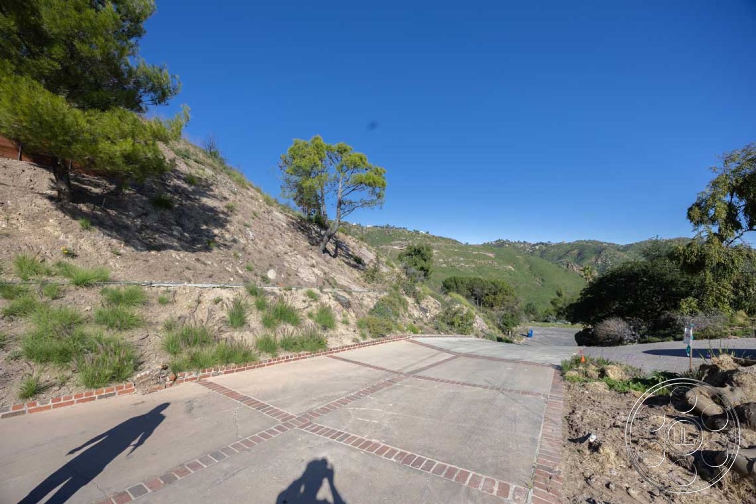 Modern 56 - concrete driveway, tropical trees, landscaped garden area, sun-drenched exterior, concrete surface texture, desert climate setting, brick border edging, natural vegetation surrounding, mountainous landscape view, asphalt access road, sloped hillside terrain, gravel driveway offshoot