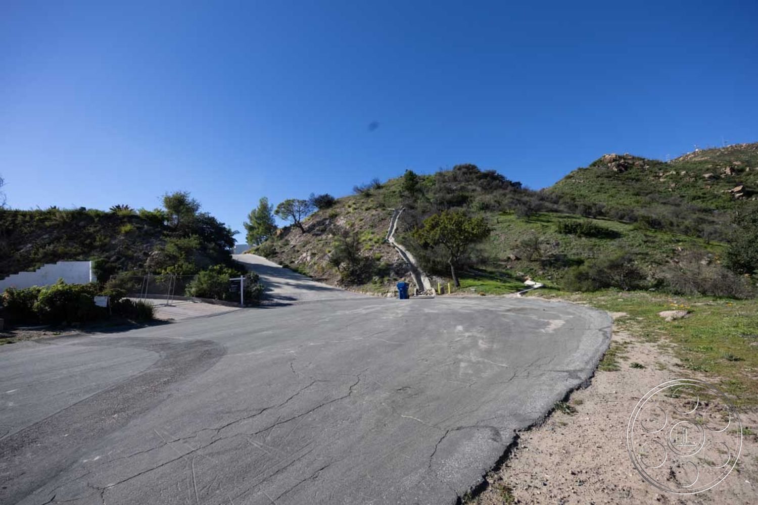 Modern 56 - exterior, curved driveway, clear blue sky, rural setting, undulating terrain, asphalt road surface, natural hillside landscape, vegetated slope, urban development site, mountain backing, seasonal greenery, scenic road, roadside vegetation
