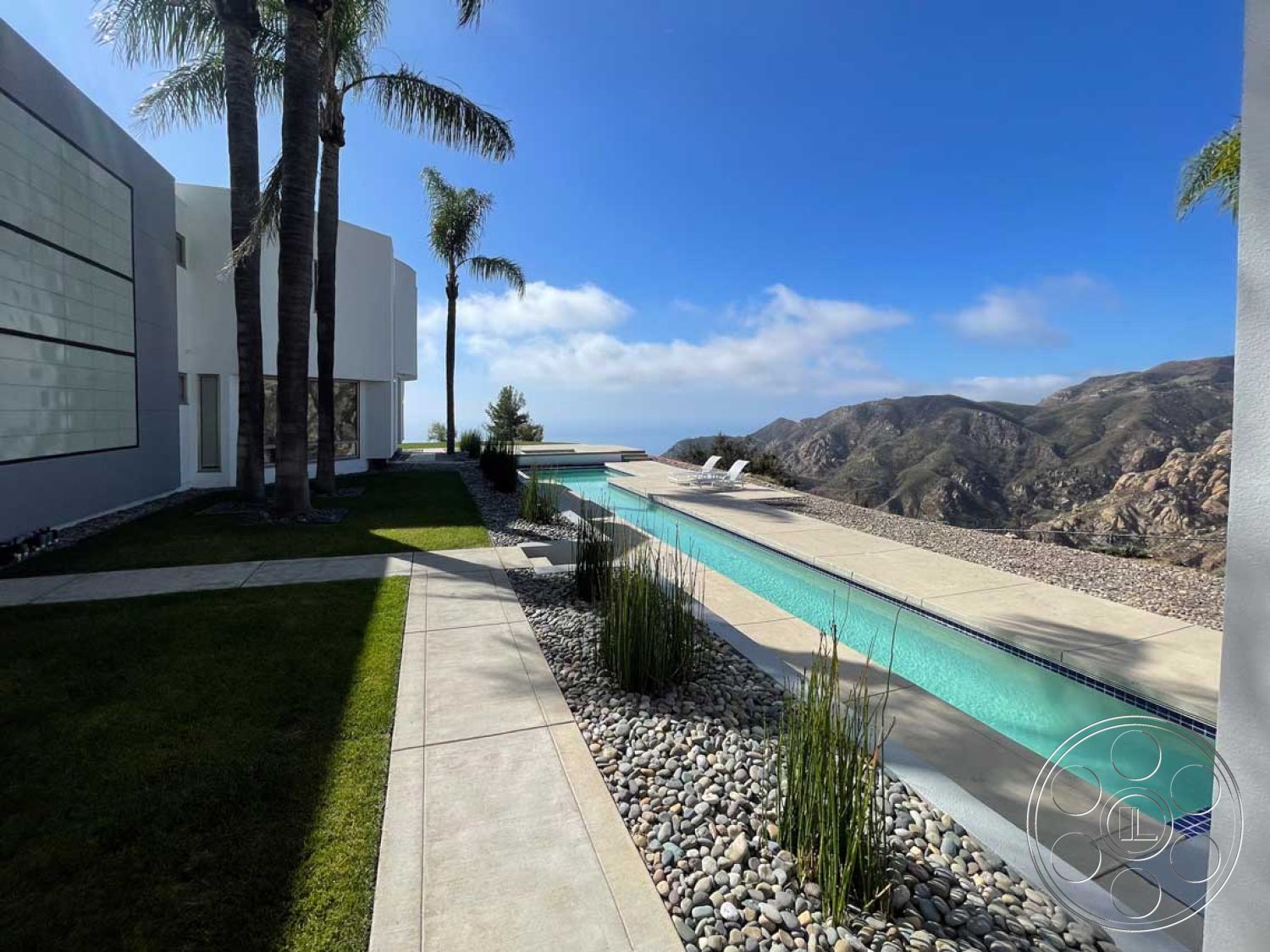 Modern 56 - natural light, exterior, floor-to-ceiling windows, modern architectural style, concrete pool deck, poured concrete pathway, gray pavers, open-air layout, mountain view setting, smooth stucco wall finish, palm tree landscaping, rectangular infinity pool, textured concrete finish, serene outdoor environment, landscaped backyard with grass, river rock garden bed, outdoor relaxation area with lounge chairs, slate stone border around pool