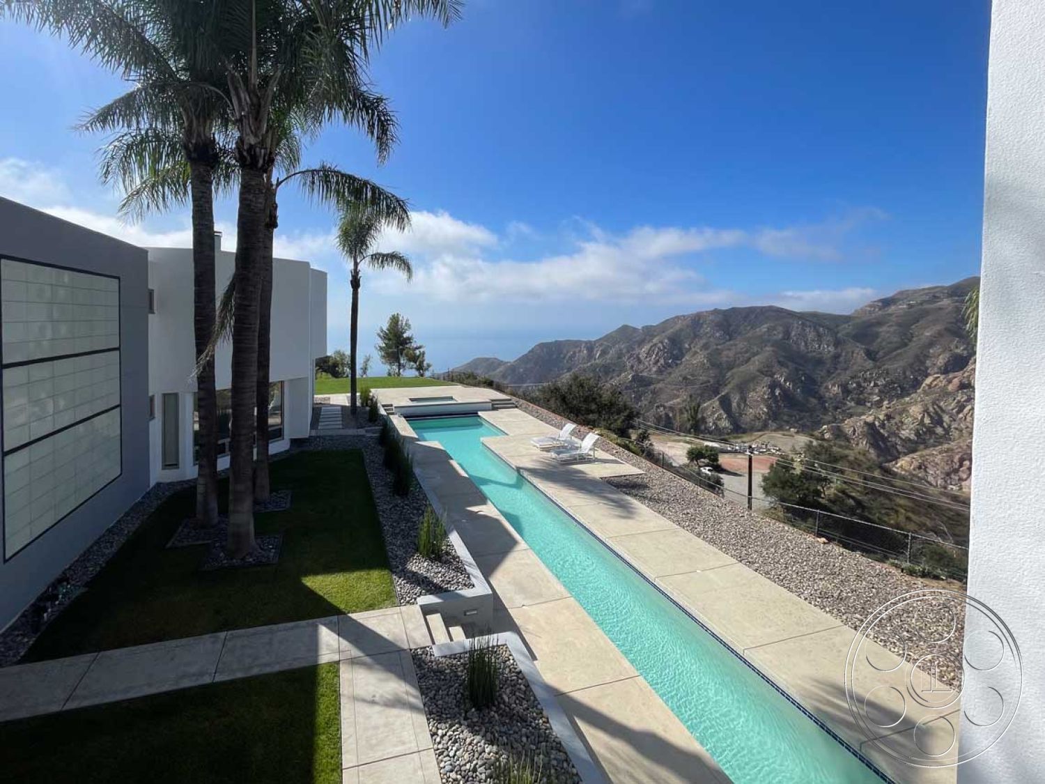 Modern 56 - palm trees, sliding glass doors, floor-to-ceiling windows, outdoor lounge area, modern architectural style, gravel landscaping, landscaped garden area, concrete patio floor, rectangular infinity pool, minimalist facade, desert mountain view, exposed concrete retaining wall, smooth white stucco exterior, paver stone pool deck, stone staircase, curved exterior walkway