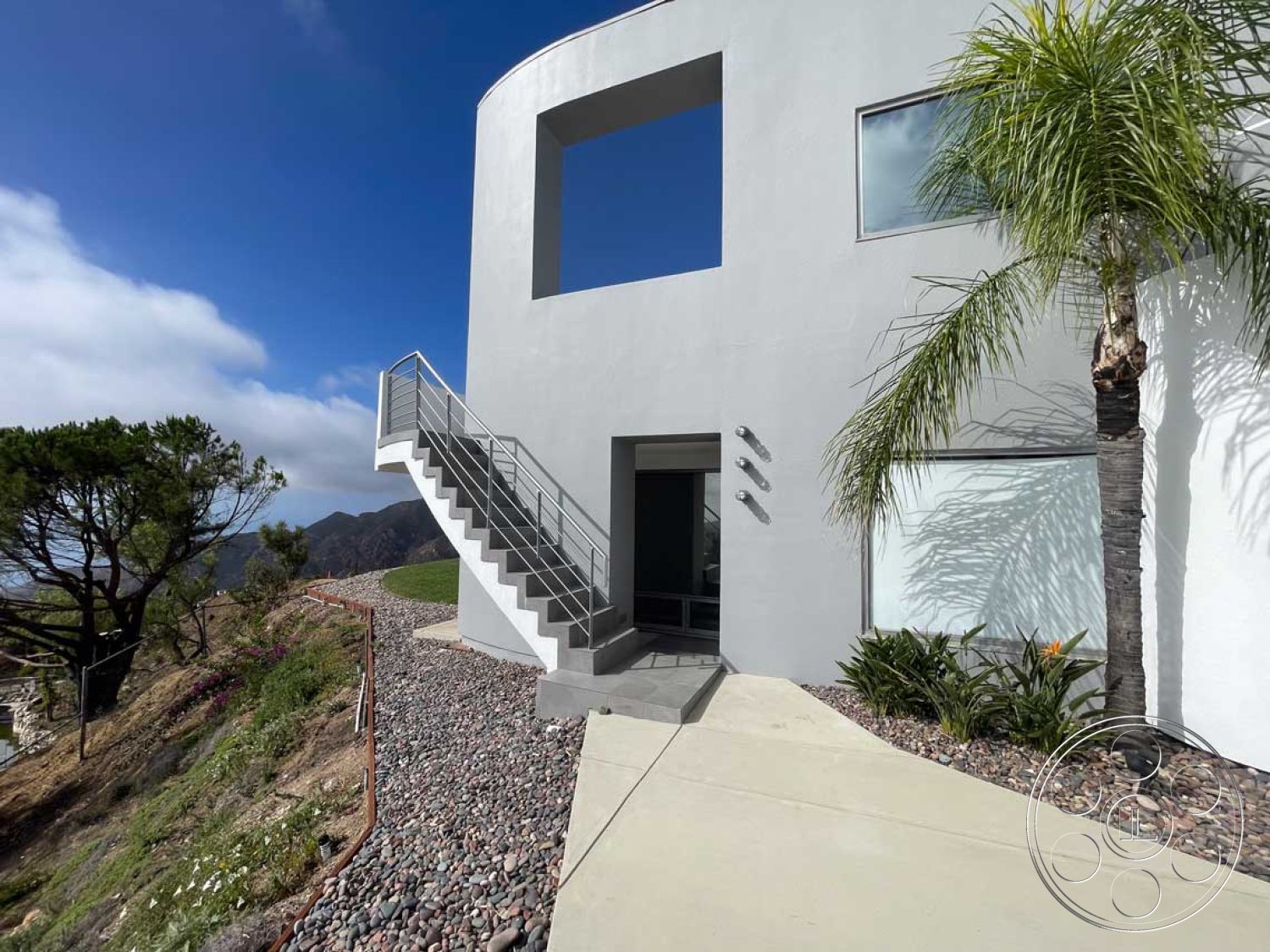 Modern 56 - minimalist design elements, floor-to-ceiling windows, modern architectural style, metal railing staircase, outdoor living area, smooth concrete surfaces, mountain view setting, stucco exterior walls, concrete patio floor, palm trees landscaping, natural light abundance, landscaped garden path, exterior lighting fixtures, pebble stone landscaping, curved architectural design, open floor plan connectivity