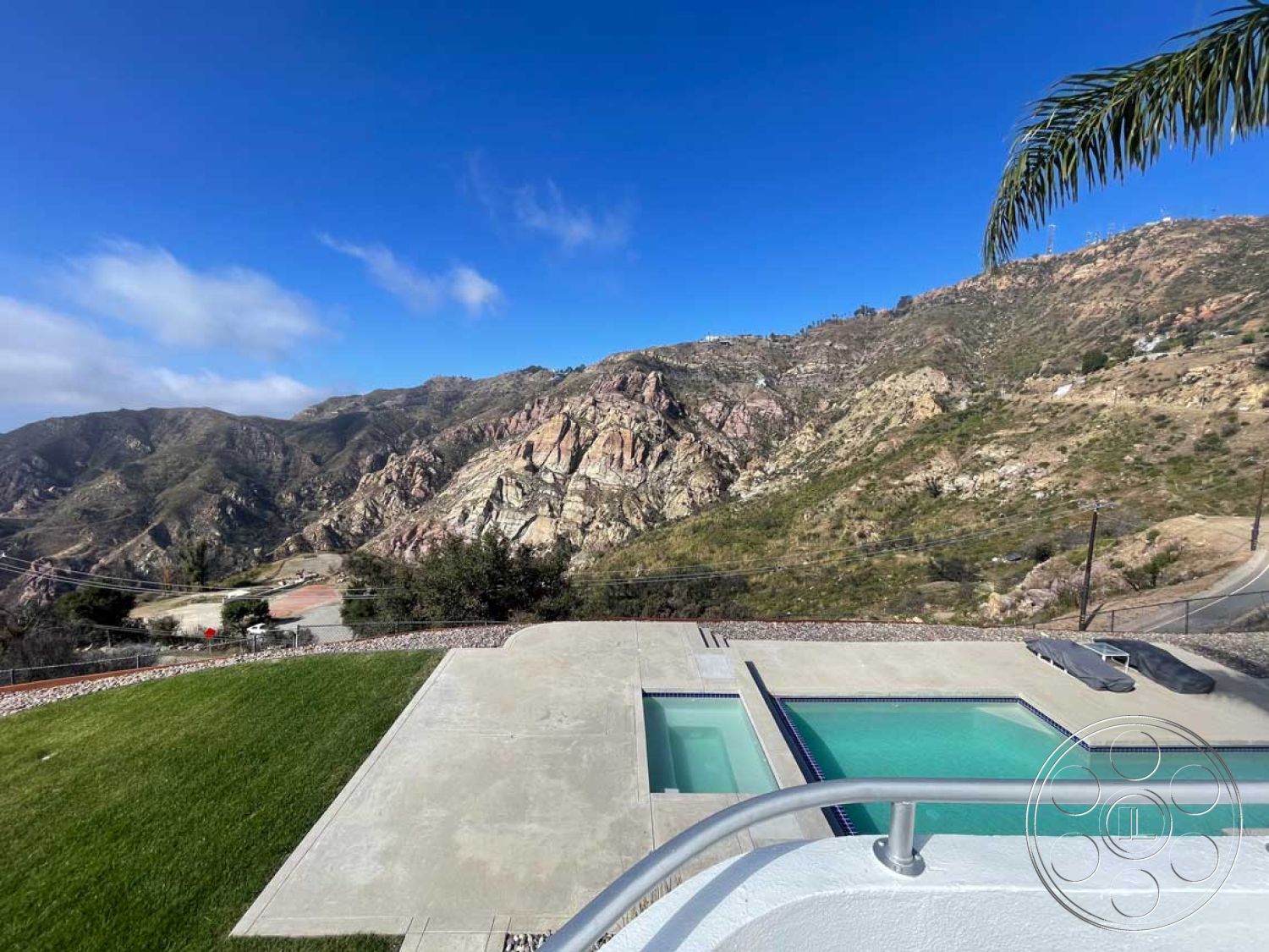 Modern 56 - exterior, landscaped garden, sloped terrain, concrete decking, lush green lawn, mountain view, clear blue sky, infinity pool, modern outdoor lounge area, covered outdoor seating area, rectangular concrete pool border, concrete pool patio flooring, surrounding rocky landscape, minimalist pool design, scenic outdoor environment