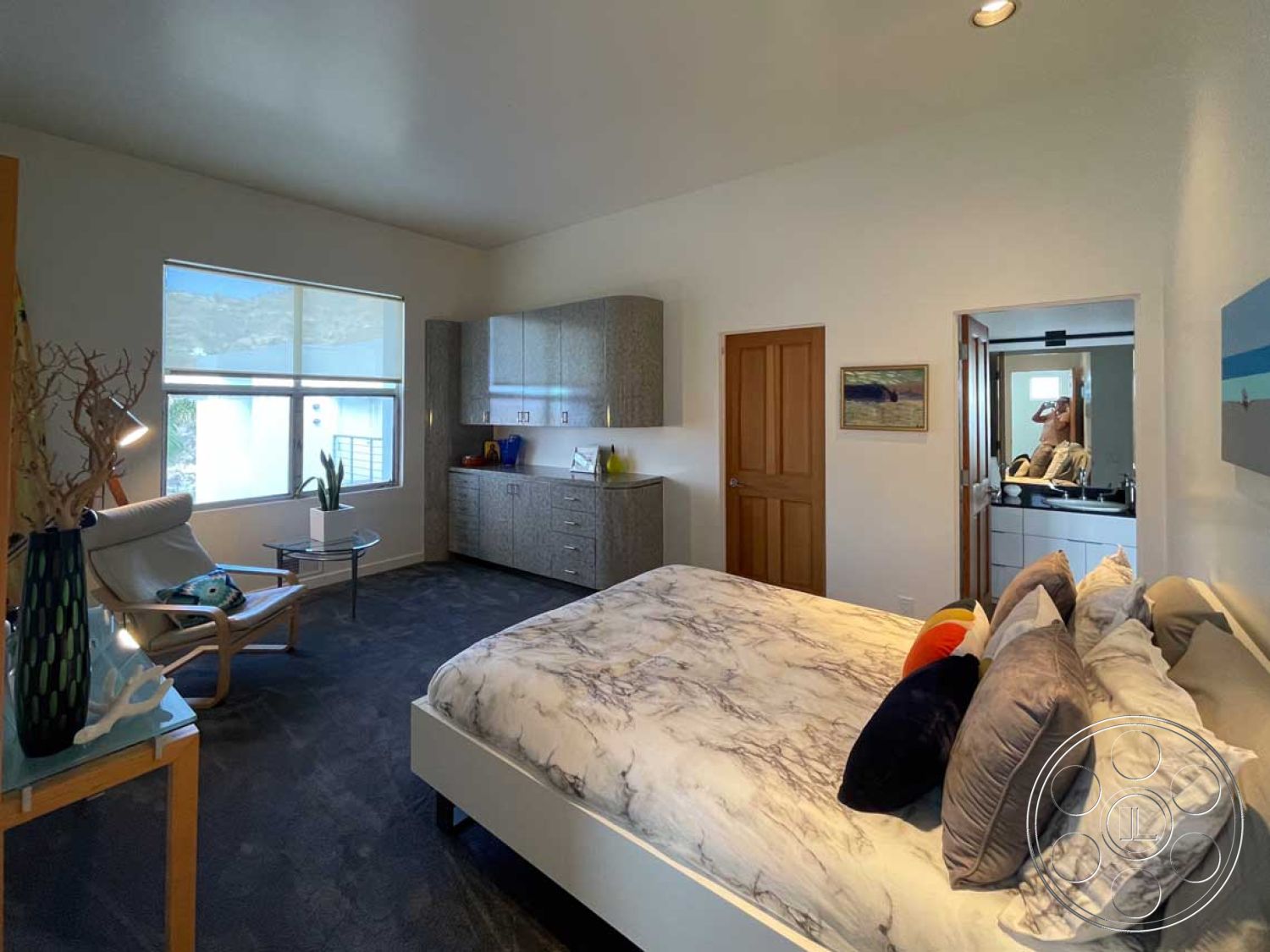 Modern 56 - neutral color palette with pops of color, interior bedroom with carpet flooring, modern furnishings with marble bedspread, glass windows providing natural light, wooden door leading to bathroom, built-in cabinetry with glossy finish, textured wall treatment with light-colored paint, contemporary accent chair with patterned cushion, compact side table for bedside use, decorative accessories with coastal theme, high ceilings enhancing spacious feel, plush pillows for comfort on the bed, artwork with abstract ocean scene, functional layout with accessible design, efficient use of space for storage, green plant accents for freshness