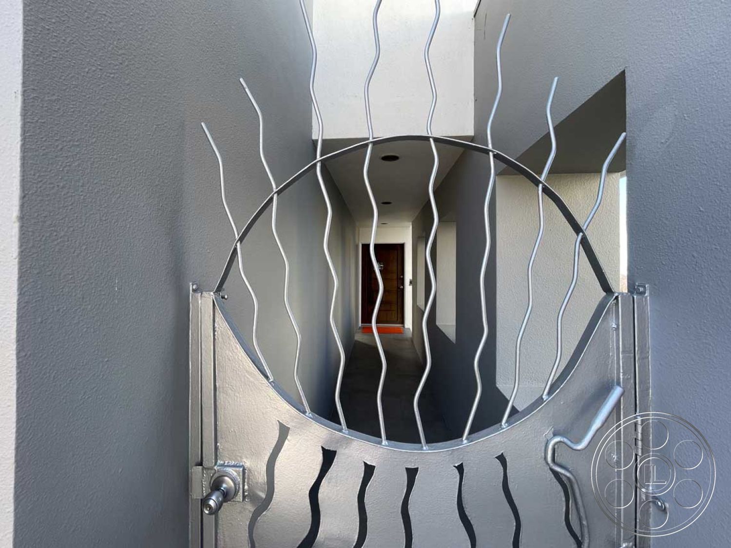 Modern 56 - neutral color palette, modern architectural style, arched entryway, wooden front door, residential exterior, textured plaster wall, exterior walkway, exposed ceiling with recessed lighting, metal decorative gate, gray cement wall, wavy metal railing, symmetrical pathway design, minimalist home entrance, interior view through gate, emphasis on lines and shapes