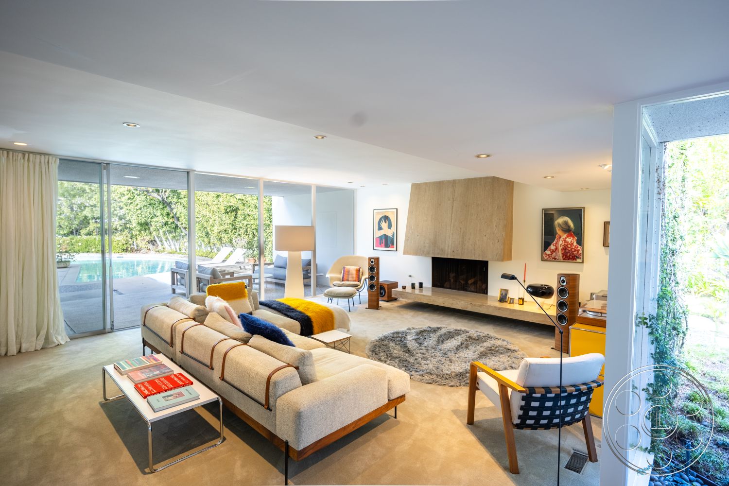 MCM 27 - mid-century modern furniture, contemporary lighting fixtures, interior living room with concrete flooring, neutral color palette with pops of color, exposed wood ceiling beams in living room, floor-to-ceiling windows overlooking pool area, modern fireplace with wood veneer surround, textured area rug in circular shape, sliding glass doors to outdoor pool, light-colored curtains for natural light, minimalist coffee table with metal frame, yellow accent throw pillows, artwork on white painted walls, indoor plants near glass partition, open concept design with seamless flow, cozy reading nook with modern armchair, swimming pool view from living room