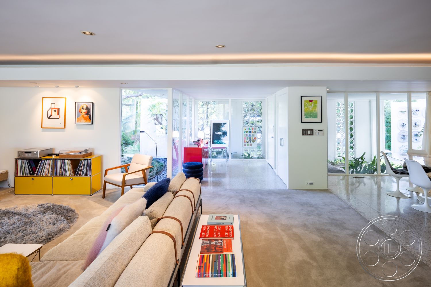 MCM 27 - indoor plants, polished concrete flooring, floor-to-ceiling windows, mid-century modern furniture, light gray sectional sofa, glass partition walls, minimalist design aesthetic, white painted walls, open concept living area, contemporary art wall decor, exposed ceiling lights, textured wool area rug, natural light filled space, access to outdoor patio, yellow wood media console, soft earth tone furnishings, modern armchair with wooden legs, living room entertainment area, vertical garden wall feature