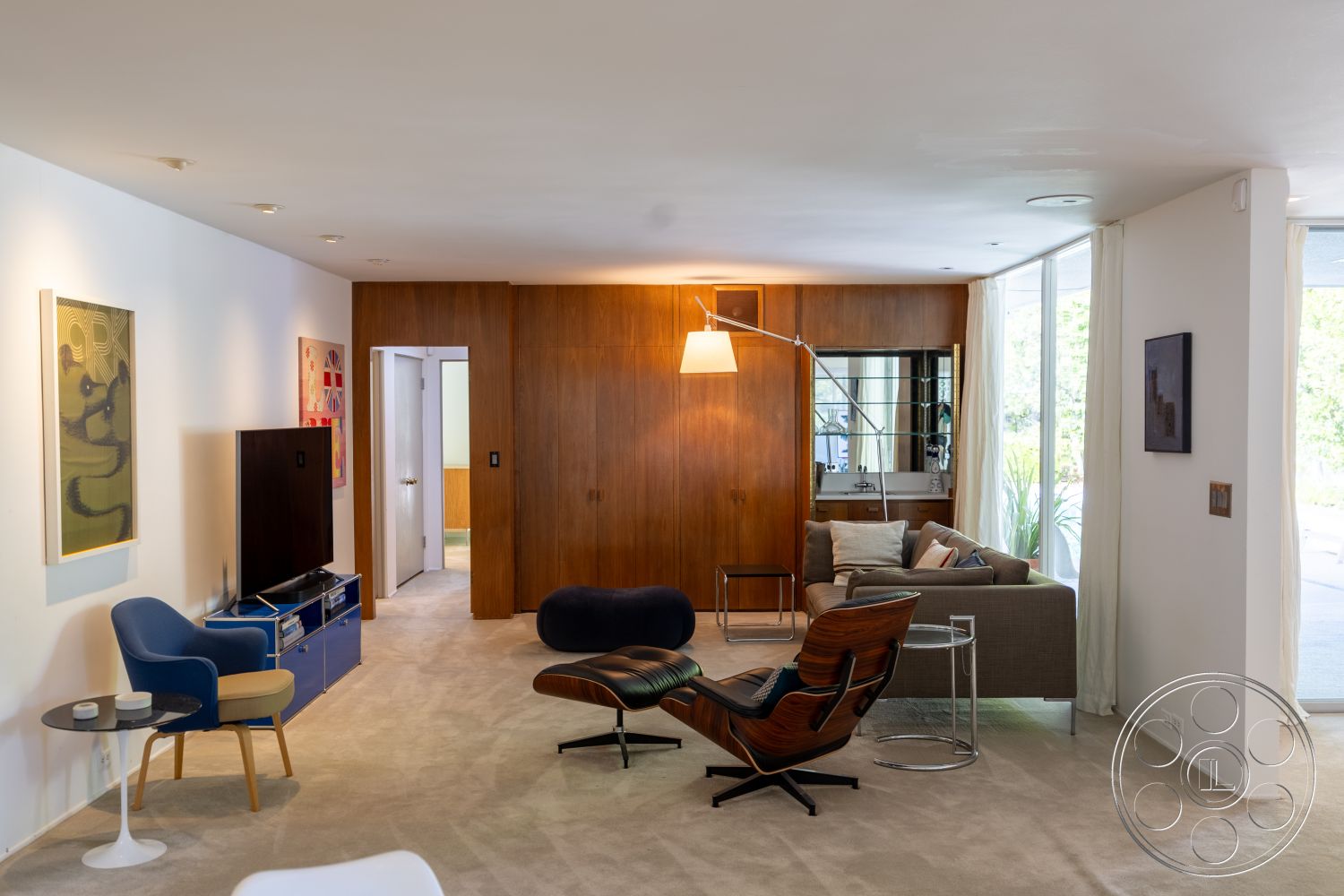 MCM 27 - interior, mid-century modern living room, carpet flooring, beige carpet living room floors, exposed wood paneling accent wall, white painted walls, light-filled space, floor-to-ceiling windows, natural light, modern furniture, blue modern chair, wooden lounge chair, decorative art pieces on wall, colorful artwork, chrome floor lamp with adjustable arm, minimalist coffee table, round glass top, open layout, seamless flow between rooms