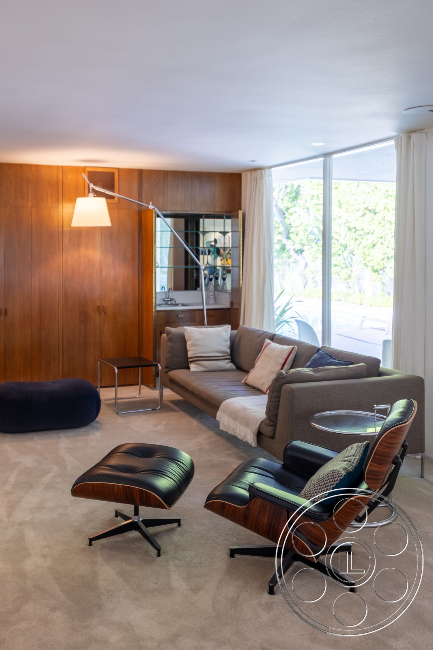 MCM 27 - neutral color palette interior, floor-to-ceiling windows interior, minimalist design elements interior, exposed wood paneling interior, natural light-filled living space interior, soft carpet flooring interior, mid-century modern design style interior, simple geometric shapes in furniture, textured fabric sofa with accent pillows interior, stylish metal side table interior, dark leather lounge chair interior, architectural floor lamp interior, stylish ottoman with wood accents interior, functional living room layout interior, lush outdoor view from interior